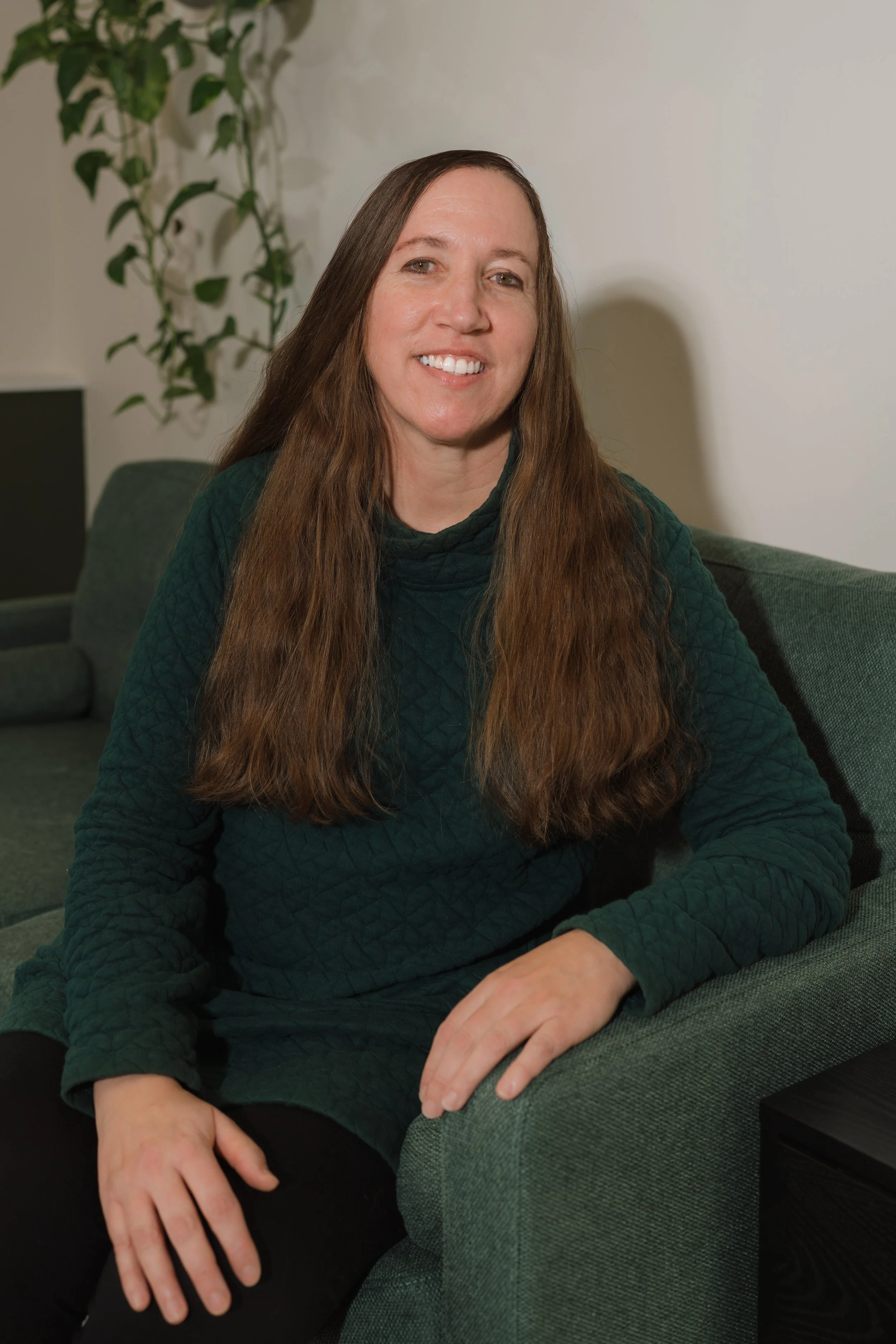 A woman with long brown hair, wearing a green quilted long sleeve top, sitting on a green couch in a room with a plant in the background.