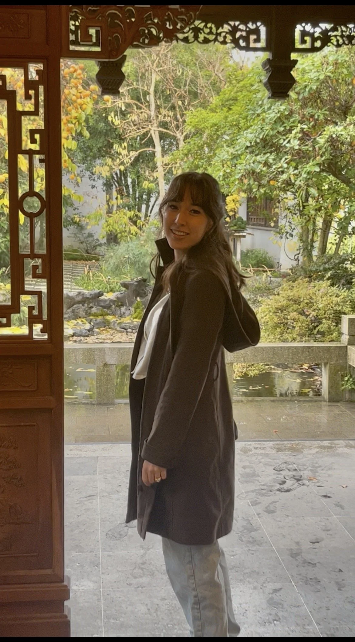 A woman with long dark hair wearing a dark coat and light pants stands near a wooden doorway with intricate carvings, smiling at the camera, with trees and yellow flowers in the background.