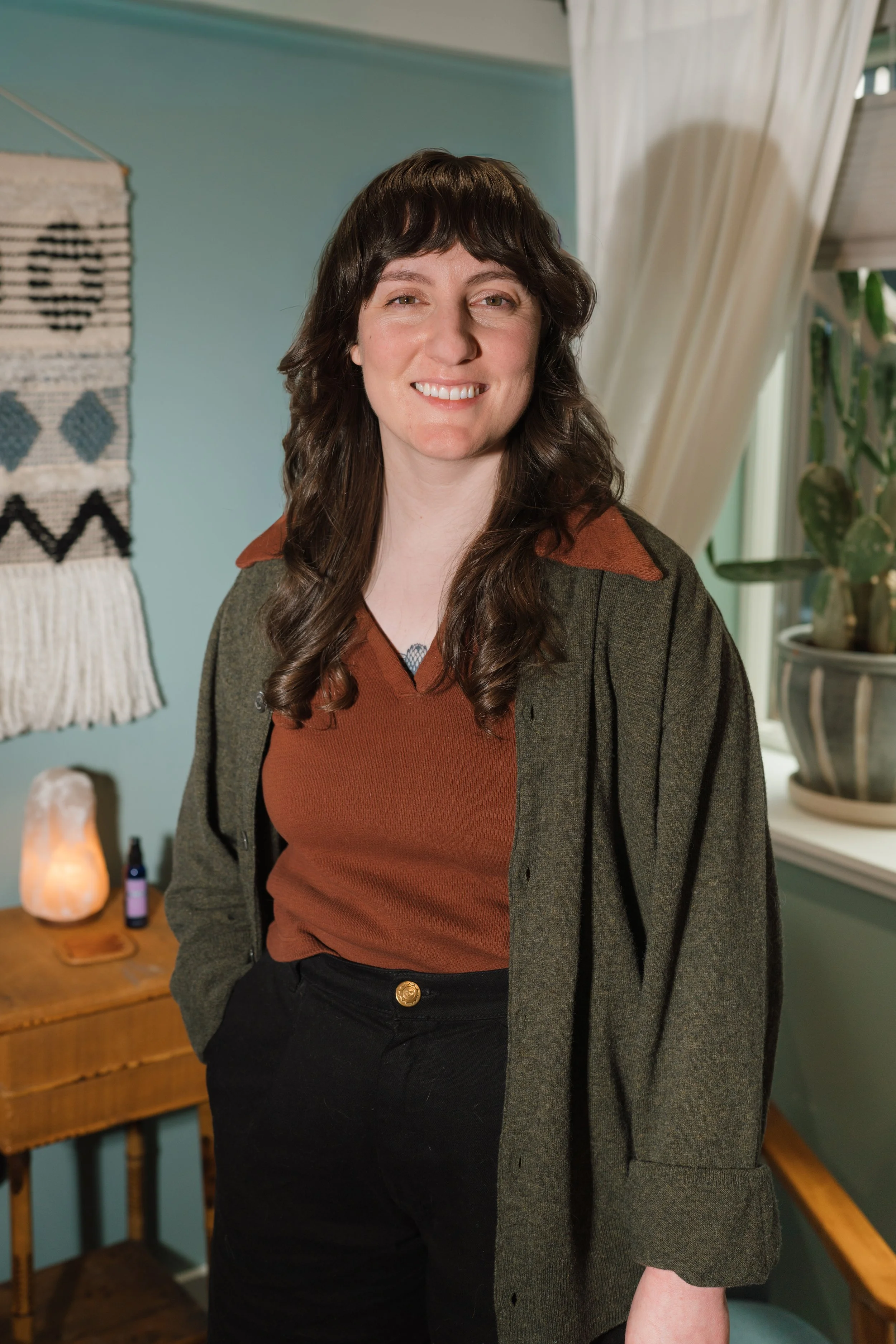 A woman with long, wavy brown hair, wearing a brown shirt with a green jacket, standing in a cozy room with teal walls, a window with white curtains, a cactus plant in a pot, a woven wall hanging, and a salt lamp on a wooden table.