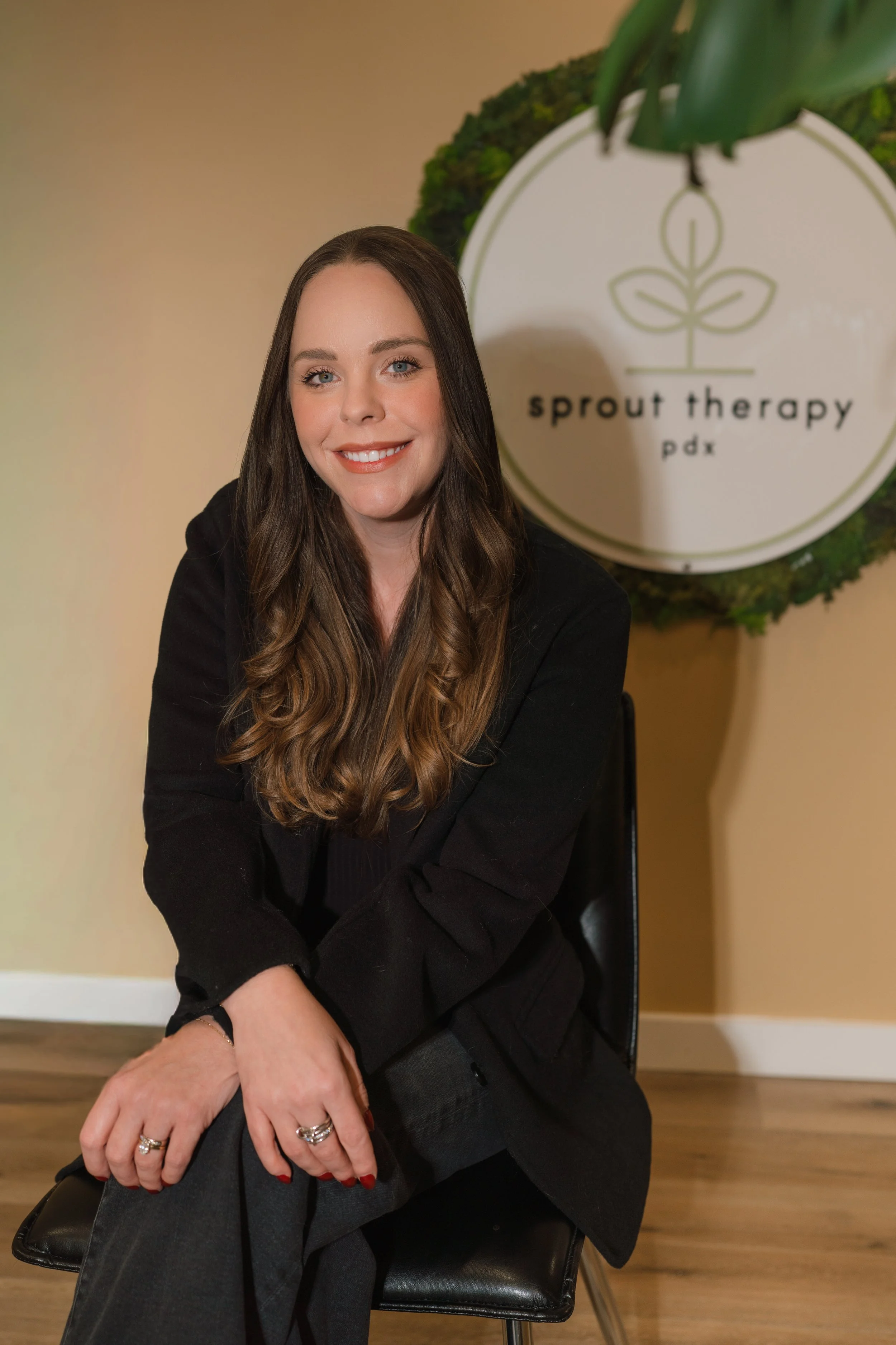 A woman with long wavy brown hair smiling, sitting on a black chair, with a sign that reads 'sprout therapy pdx' in the background.