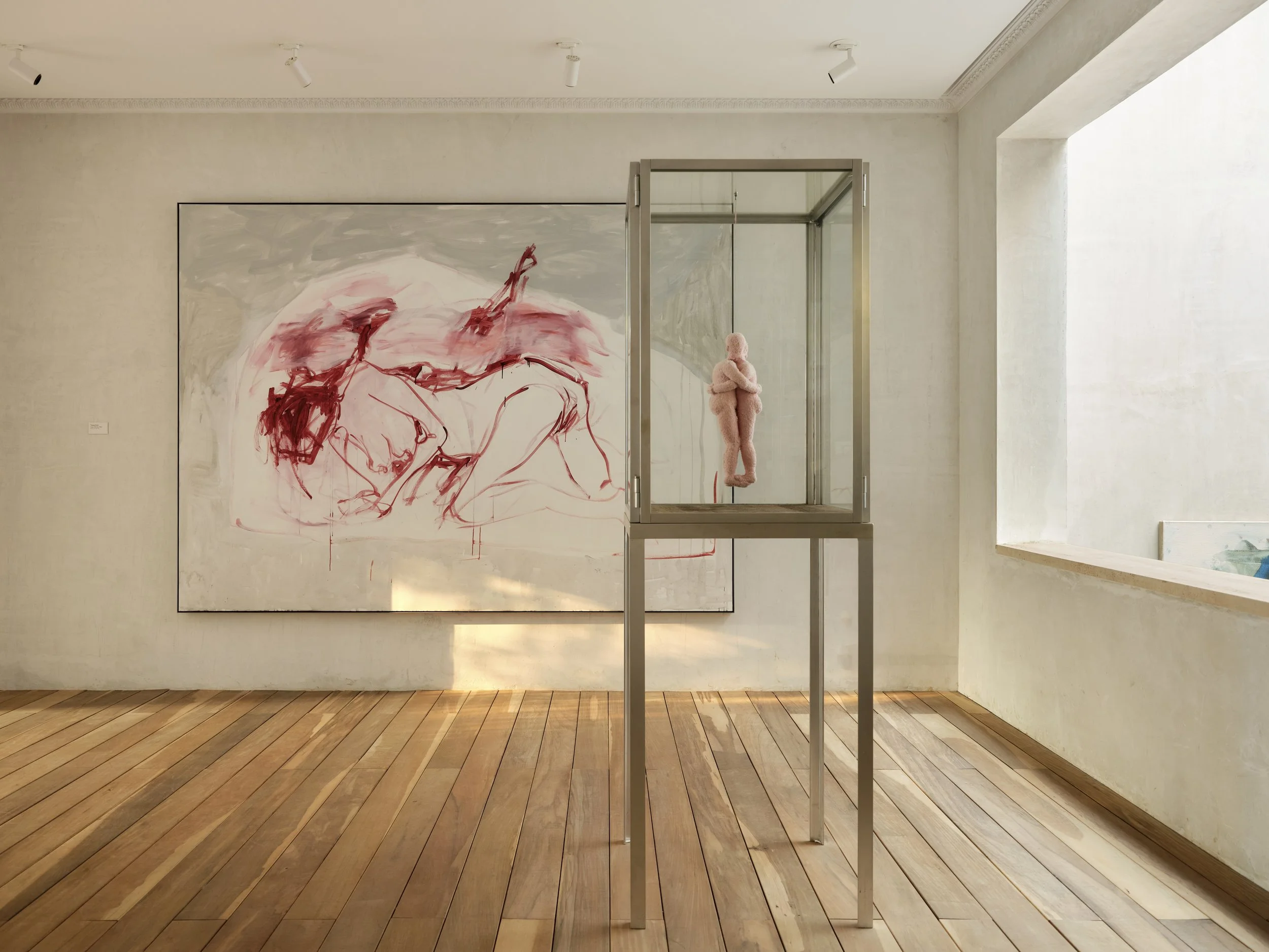 Art installation in an art gallery featuring a glass display case with a pink stuffed animal inside, a wall with a large abstract painting with red and grey tones, and wooden flooring with white walls and ceiling.