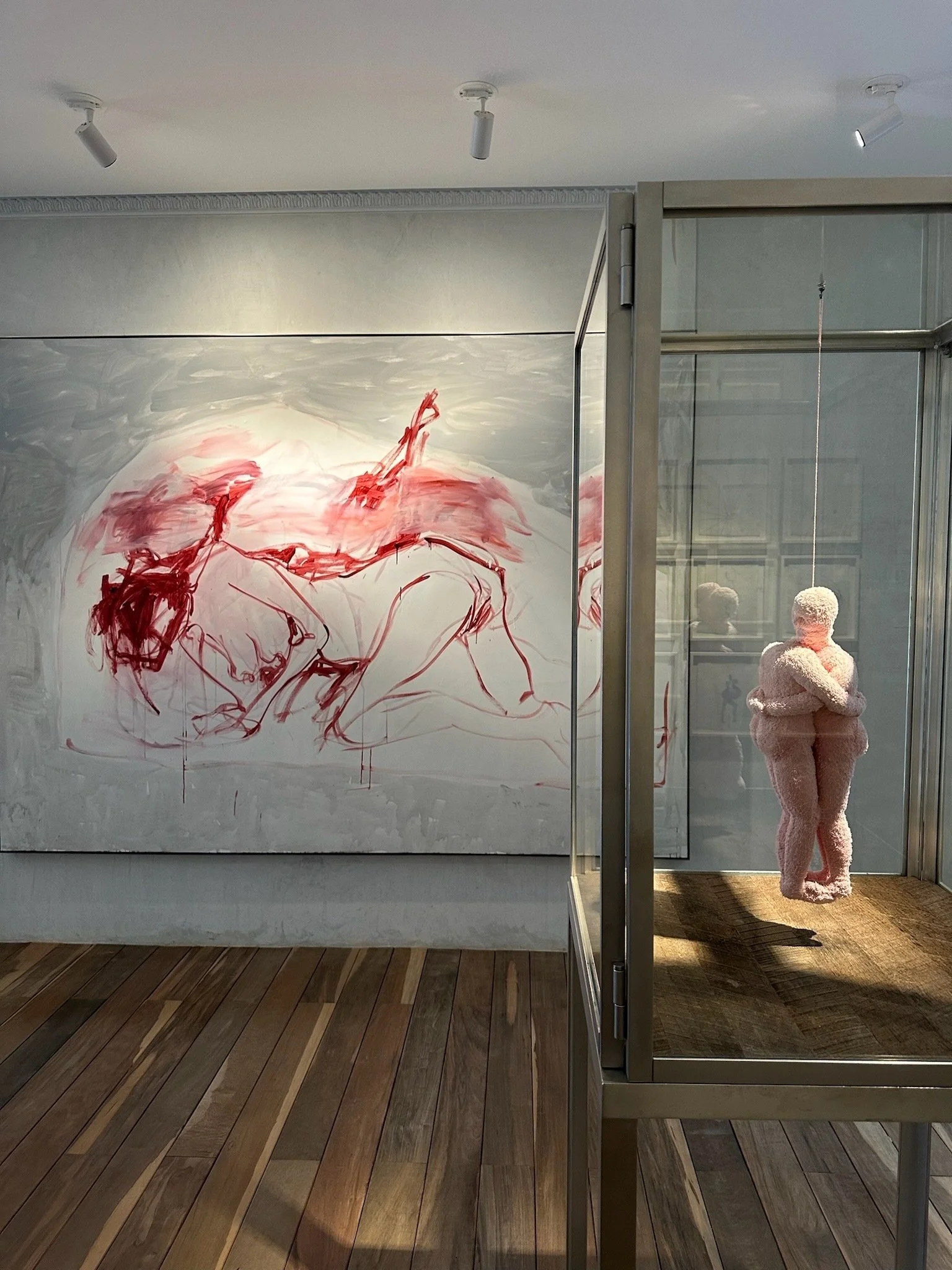Art gallery display featuring a painting of loosely sketched figures in red on a gray background and a pink, cloth-like sculpture of two people embracing inside a glass case.