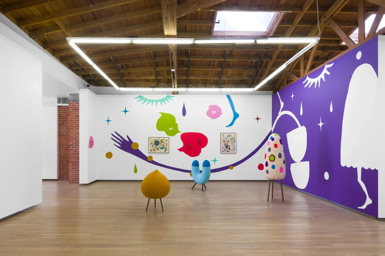 Art gallery with colorful abstract wall paintings, three unique upholstered sculptures on wooden legs, and a wooden ceiling with exposed beams.