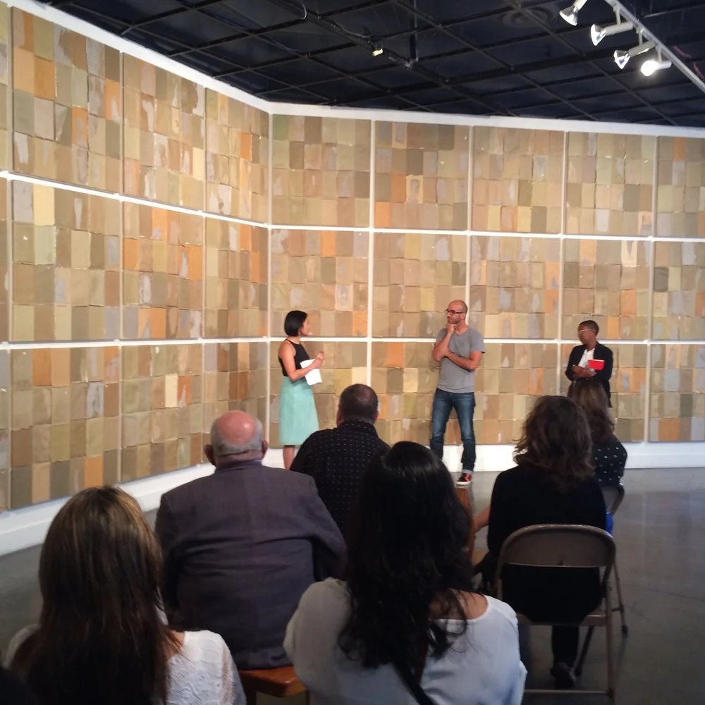 People are speaking at a panel discussion or presentation in an art gallery with a large beige tiled wall and ceiling lights.