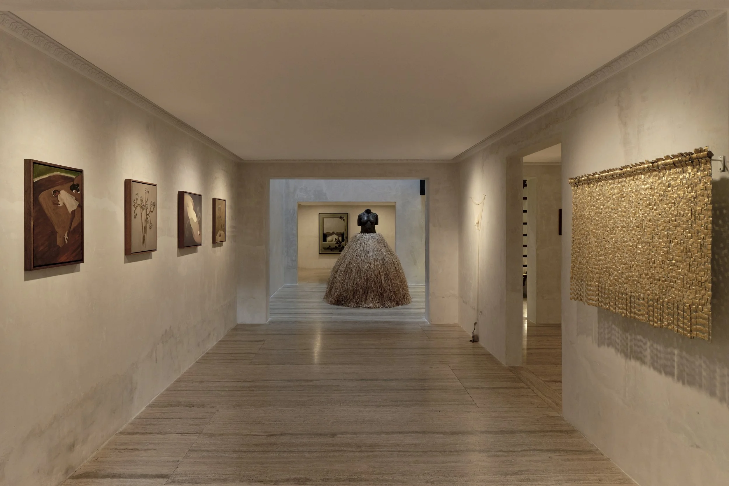 An art gallery with framed paintings on cream-colored walls, and a central sculpture resembling hair or straw, with a black dress on top, on a round pedestal.