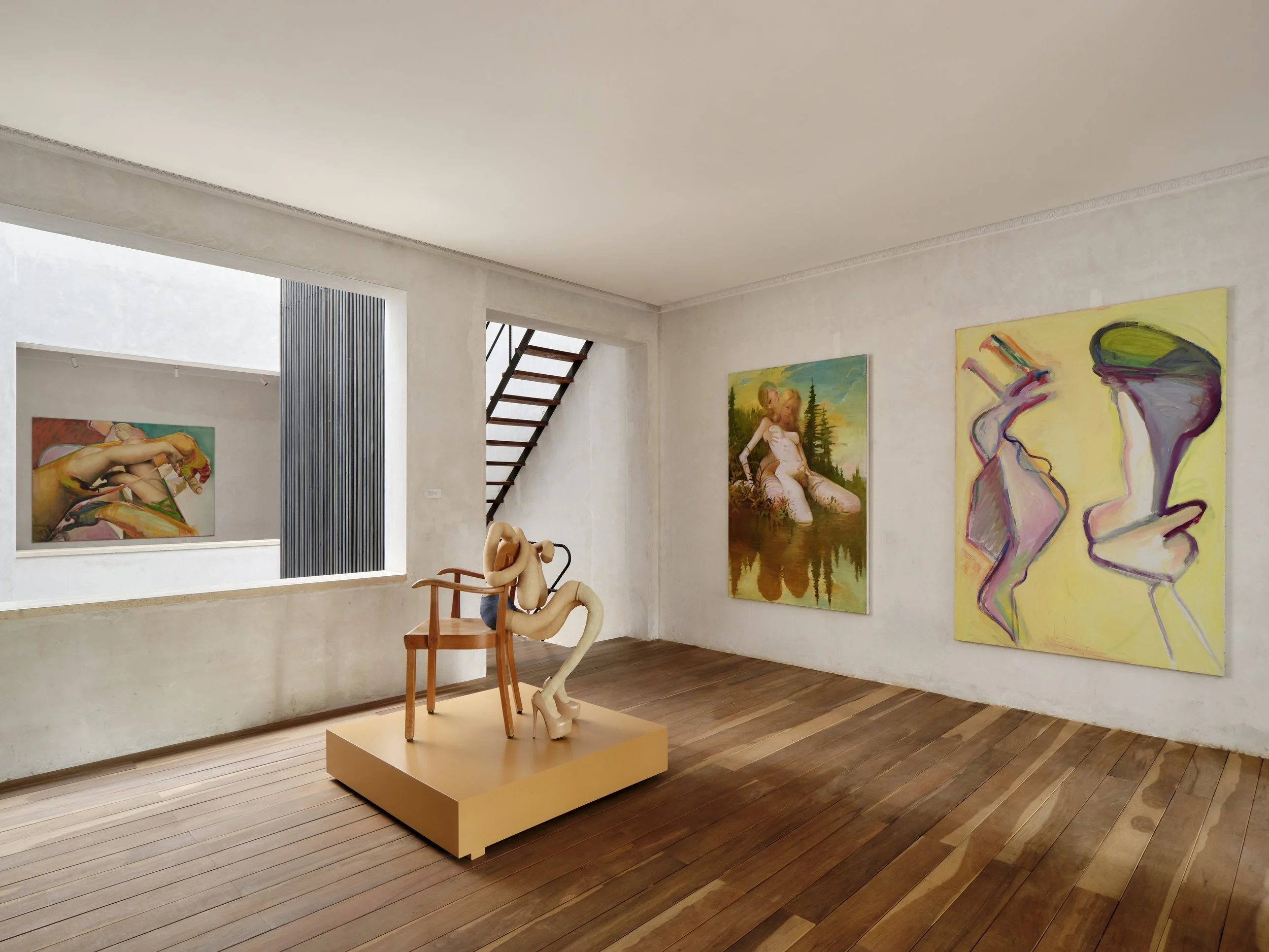 Contemporary art gallery with several colorful paintings on white walls, wooden flooring, and a sculpture of intertwined limbs on a yellow platform.