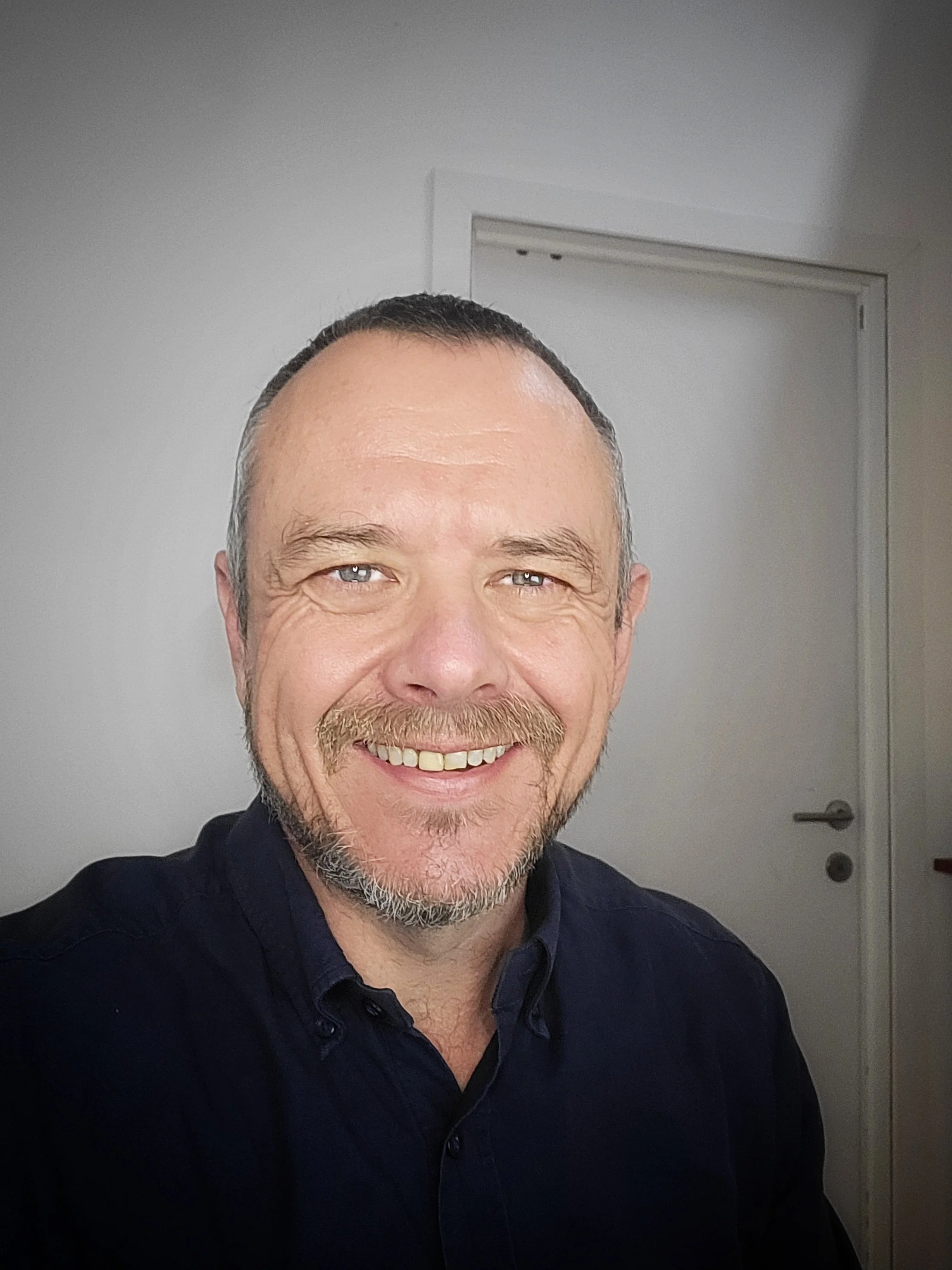Gary Bridgeman, Independent Philosopher. A middle-aged man with short dark hair, a goatee, and light-colored eyes is smiling in a photo taken indoors. He is wearing a dark collared shirt and standing in front of a plain wall and a closed door.