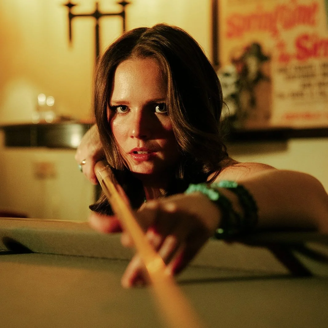 A woman with shoulder-length brown hair, looking intensely at the camera, is reaching across a surface with a stick or pencil in her hand. She wears bracelets on her arm and is in a dimly lit room with warm yellow lighting, with posters or artwork on the blurred background wall.