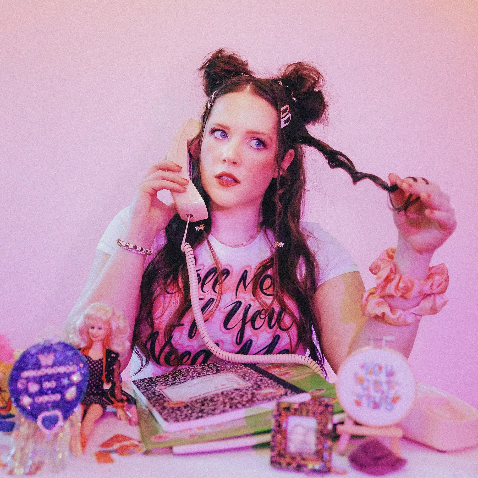 Young woman with colorful makeup and styled hair, holding a vintage pink telephone receiver to her ear, sitting at a table with toys, a diary, and a photo frame, against a pink background.