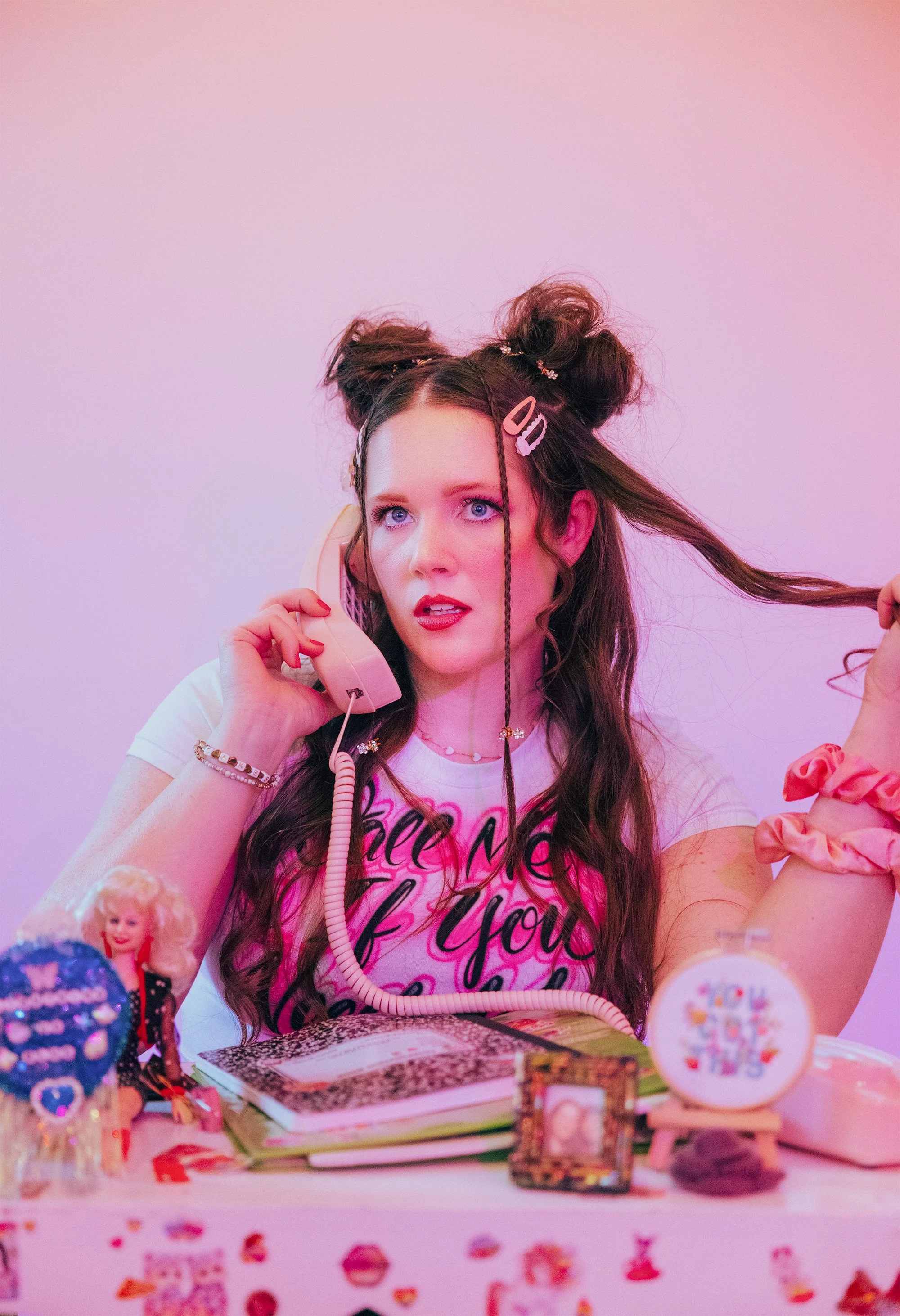 A young woman with blue eyes and long wavy brown hair styled in buns, adorned with hair clips, is speaking on a beige rotary phone. She wears a white T-shirt with pink and black text, a pink scrunchie on her wrist, and jewelry including a necklace and bracelets. The background is pink, and the table in front has various decorative items, including a doll, picture frame, and stickers.