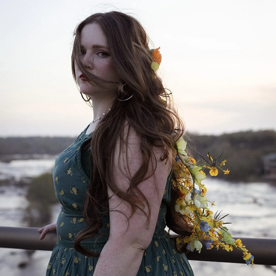 A young woman with long, wavy auburn hair and fair skin stands outdoors near a body of water during sunset. She wears a teal dress with yellow floral patterns and has a floral embellishment in her hair and on her shoulder. The background is a peaceful, natural landscape with water and distant trees.