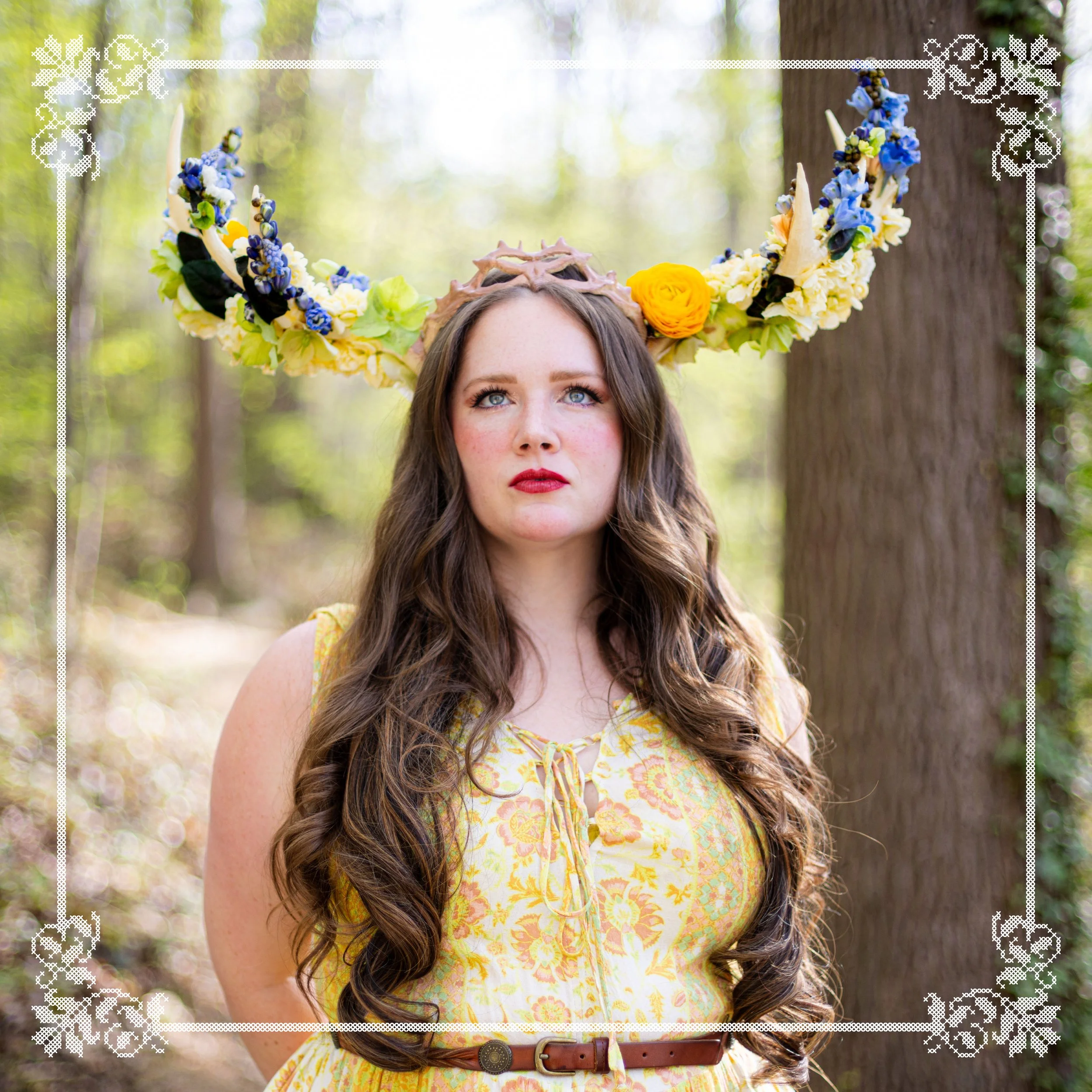A woman with long brown curly hair wearing a yellow floral dress with a lace-up neckline and a brown belt, standing outdoors in a forest with green trees in the background. She is wearing a large flower and floral crown with blue, yellow, white, and green flowers and a headpiece with a mud-like texture on her forehead.
