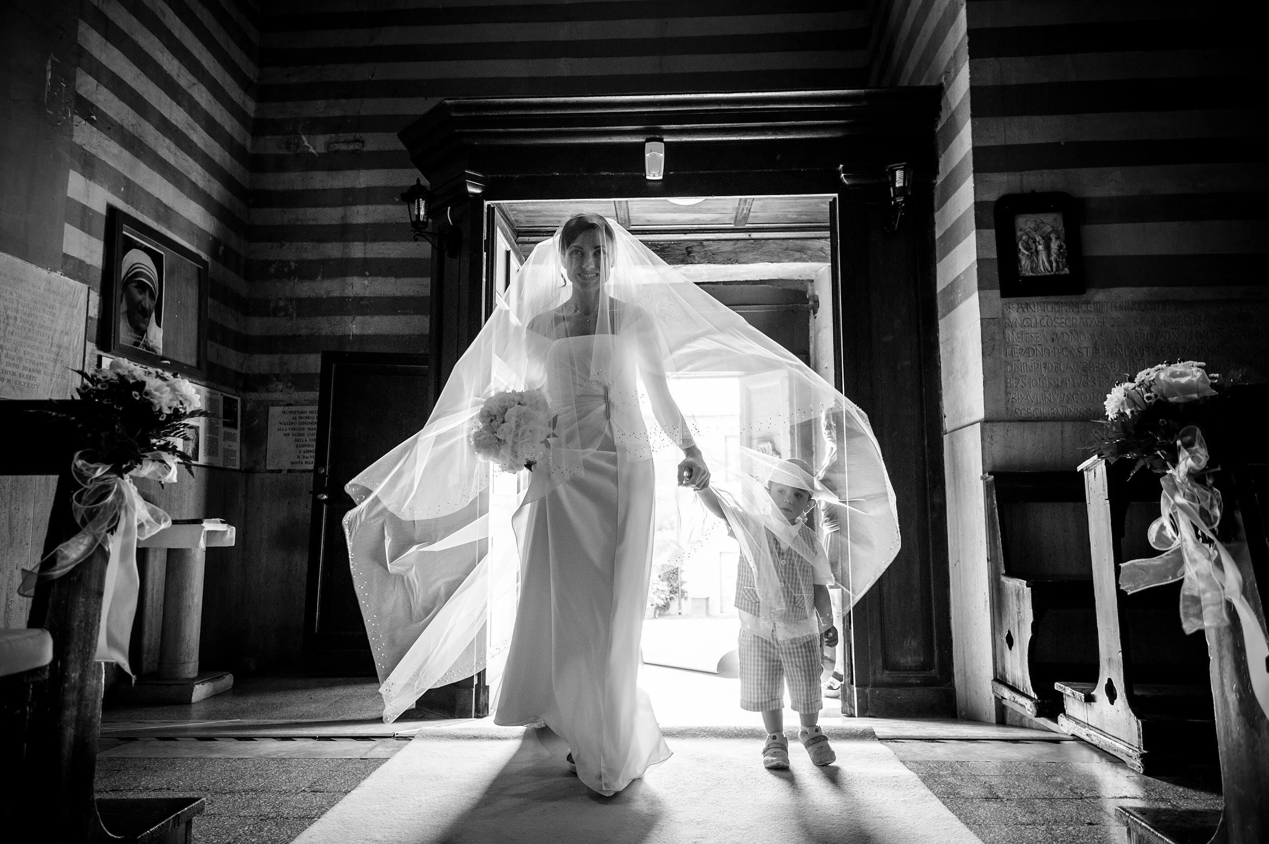 thr-bride-enters-the-church-holding-her-son-by-the-hand-big-bridal-veil-in-italy-black-and-white-wedding-photography.jpg