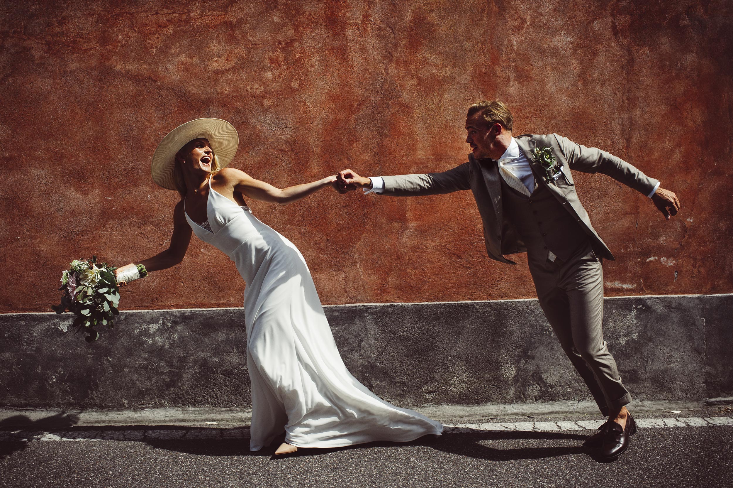 lake-como-wedding-photographer-Alessandro-Avenali-bride-with-hat-grabs-the-groom-running-against-a-red-wall.jpg