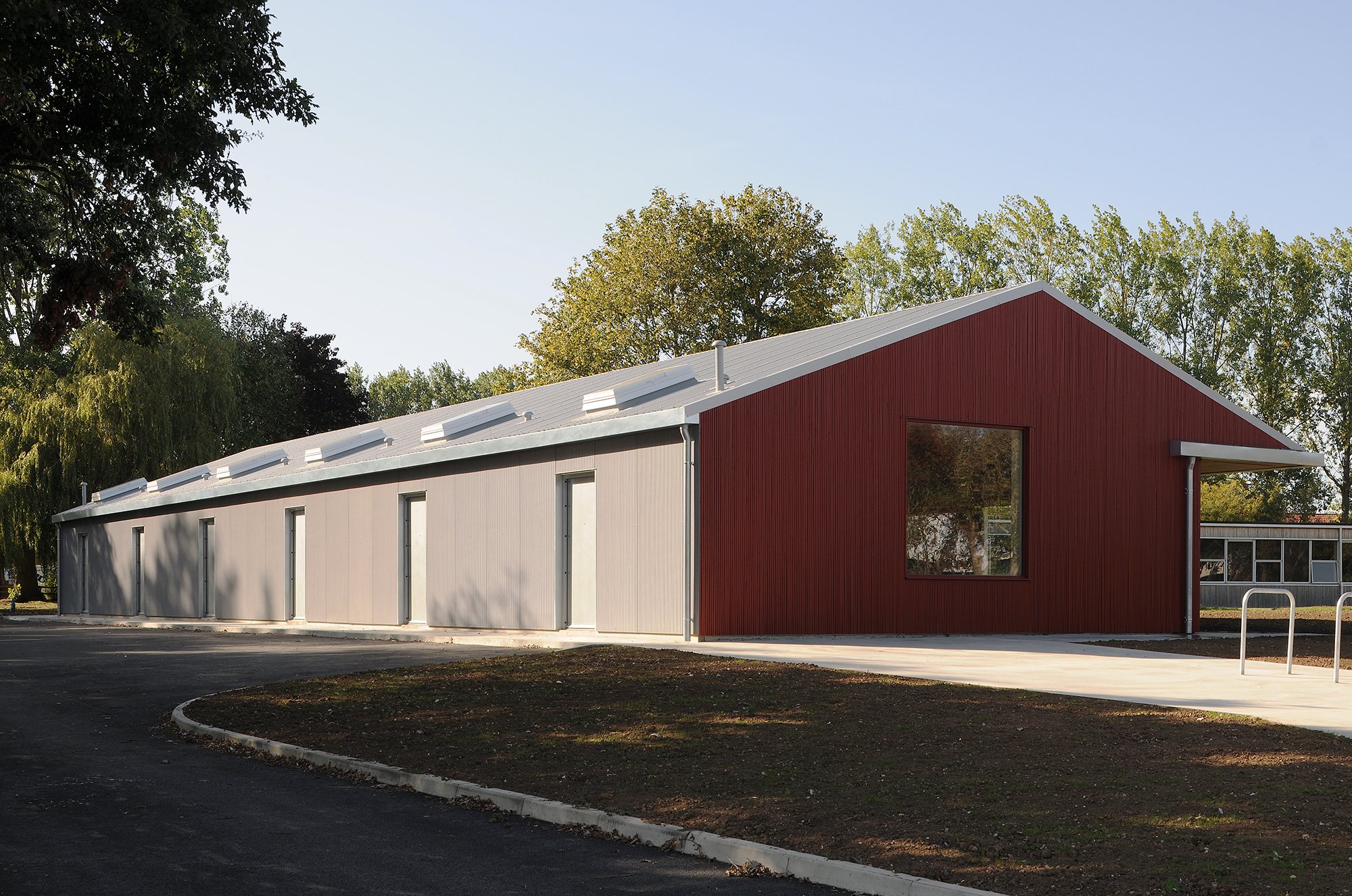 Modern building with a red and gray exterior, partial view of a large window, and a sloped roof with solar panels, surrounded by trees and a paved area.