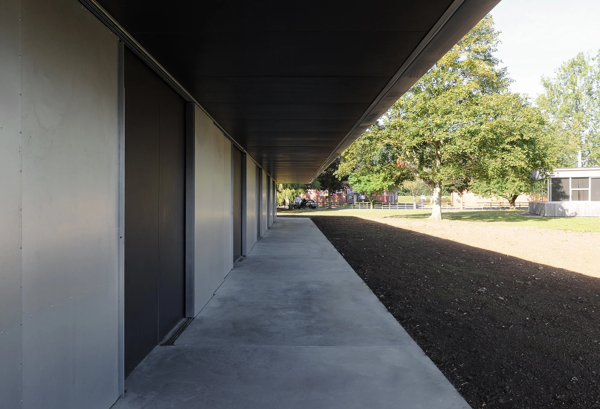 Under a modern building with a black overhang, a shaded concrete sidewalk runs along gray panel walls. In the background, green trees and a white building are visible.
