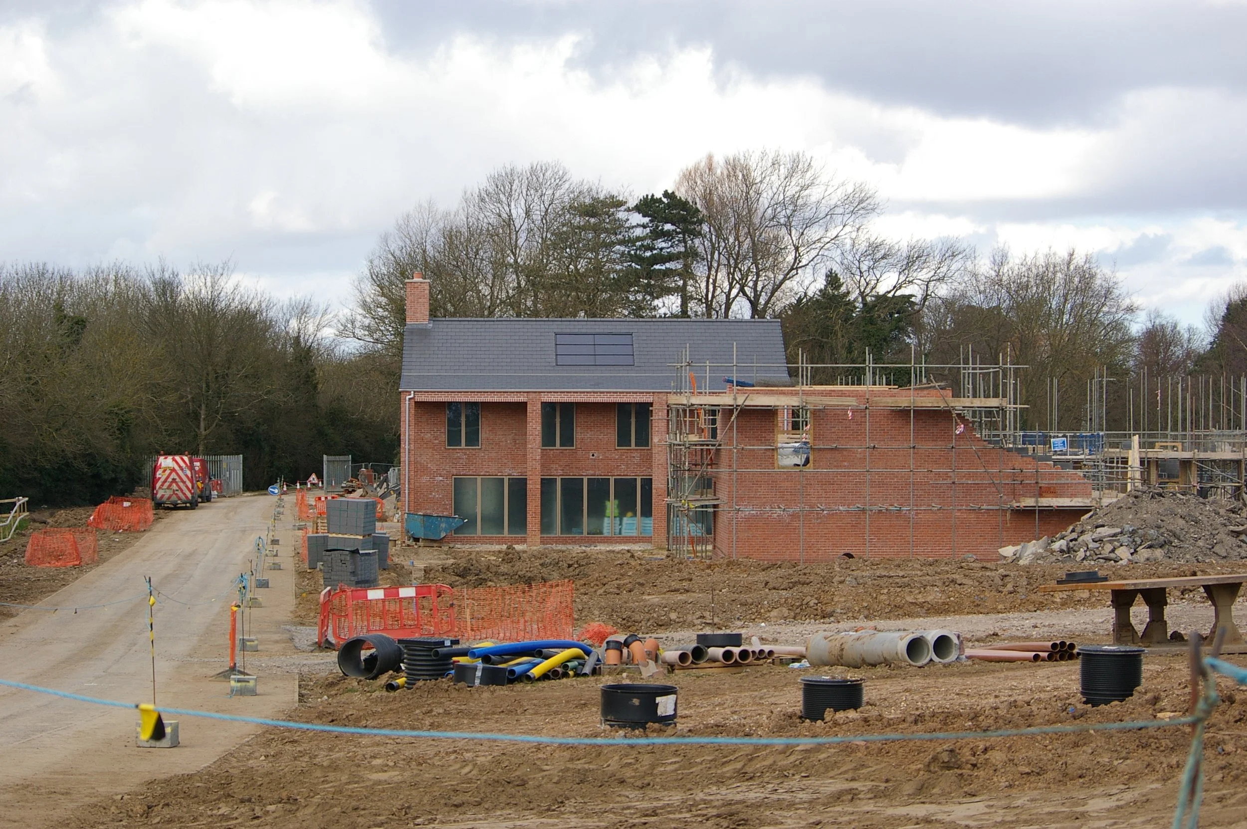 Construction site with a brick house under construction, surrounded by trees and construction materials like pipes and barriers, with cloudy sky overhead.