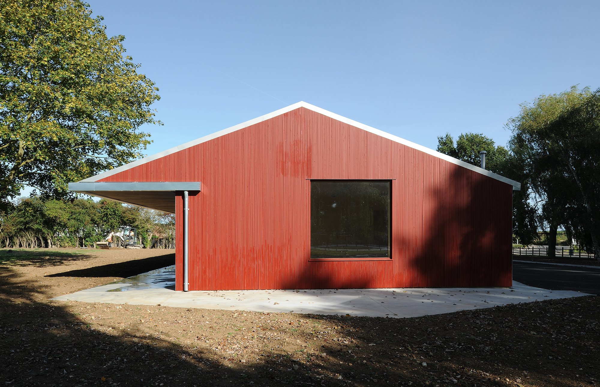 Red modern house with large window, situated on a patch of land with some trees in the background, under a clear blue sky.
