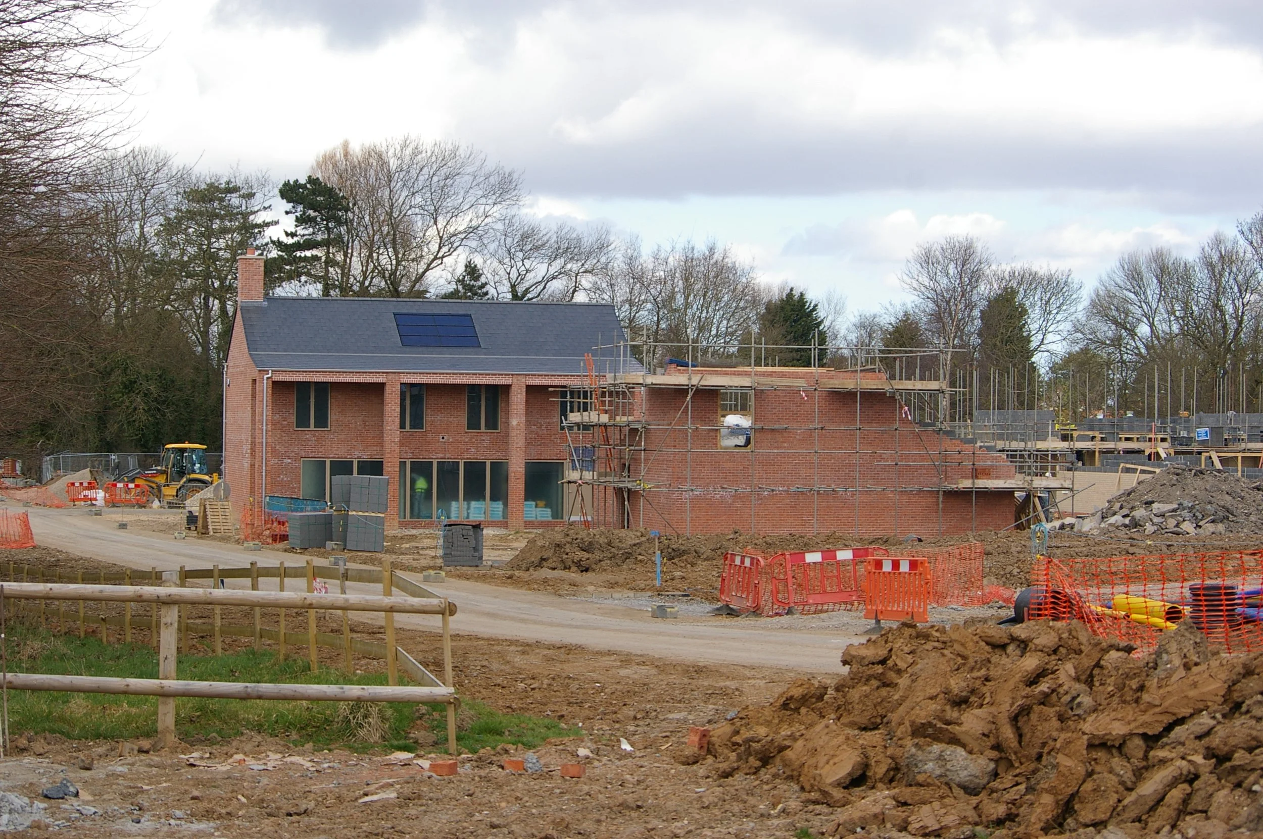 Construction site with a brick house, scaffolding, construction materials, and equipment, surrounded by trees under a cloudy sky.