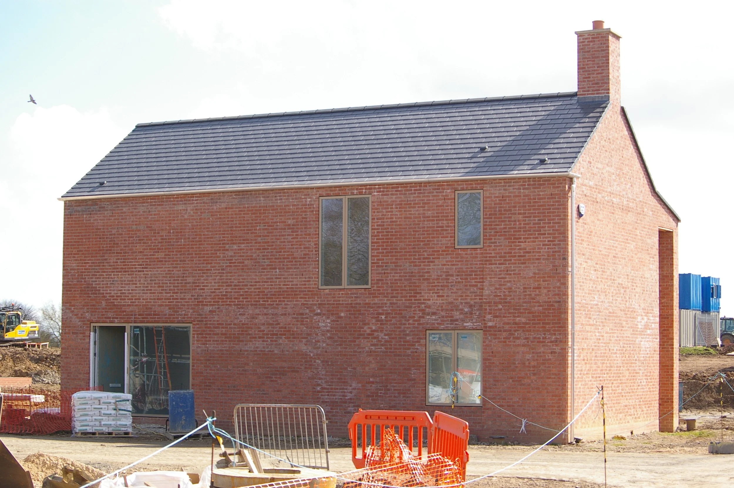 A brick house under construction with a new dark gray tiled roof, multiple windows, and a chimney. Construction materials and equipment are visible in the foreground, with safety barriers around the site.