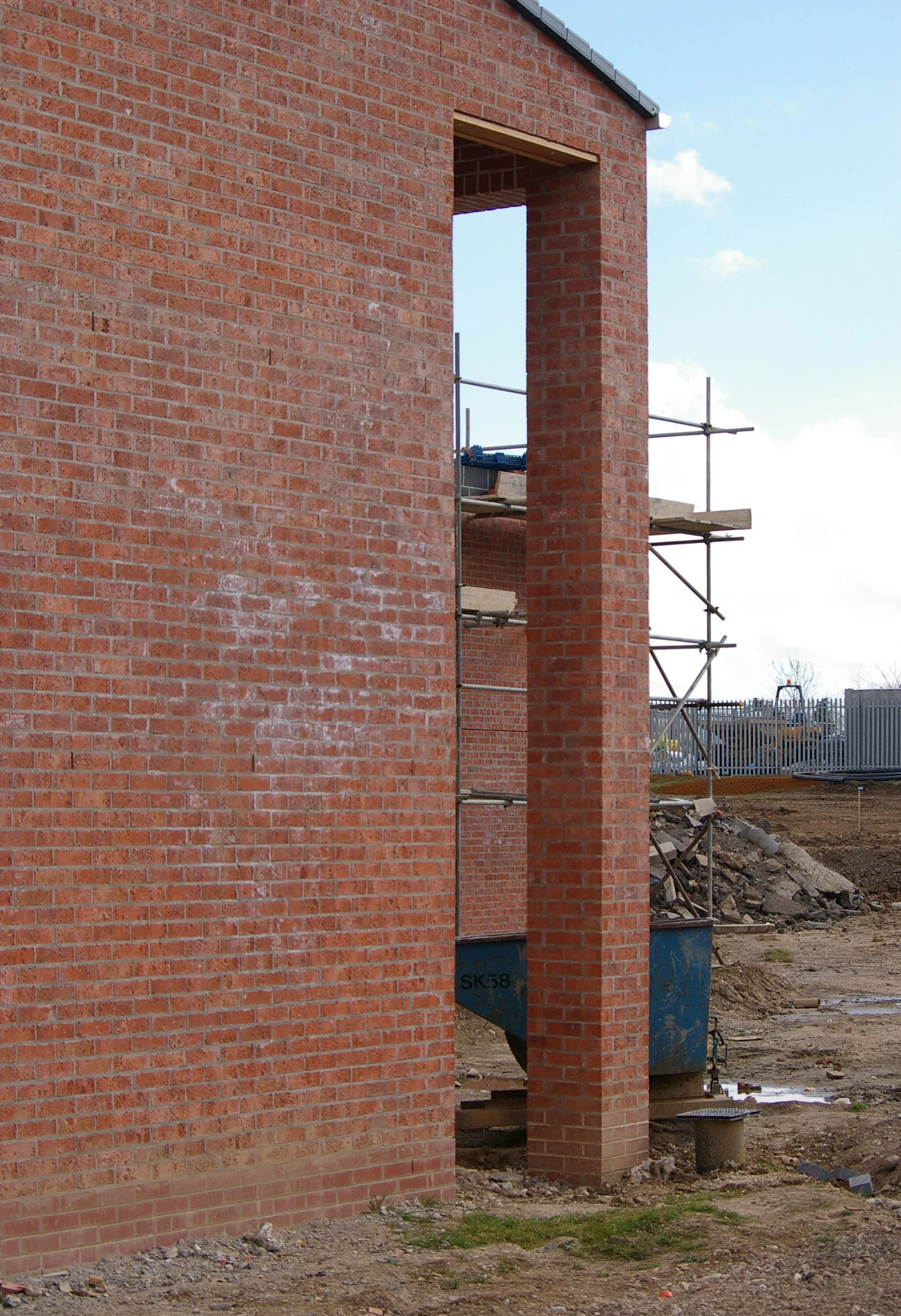 Construction site with scaffolding next to a brick wall for building a chimney or vent, with some construction materials and dirt ground visible.