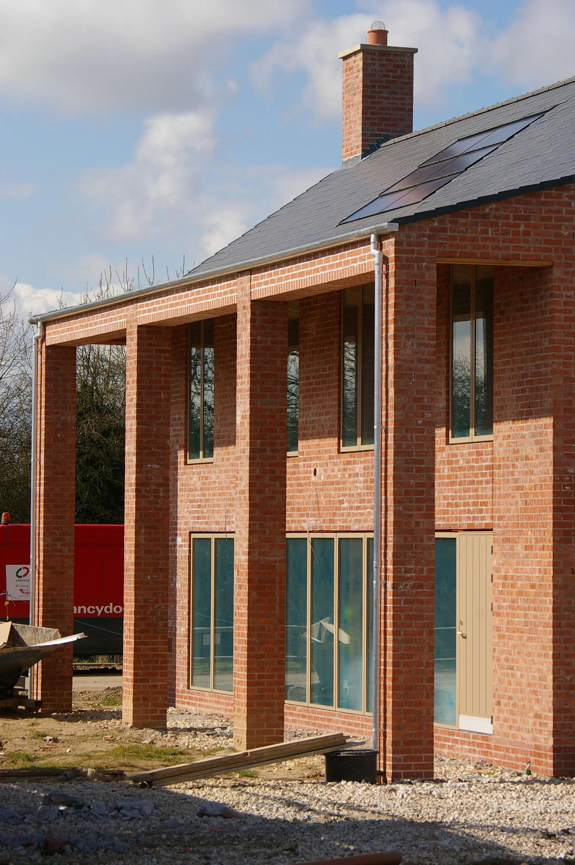 Under construction brick house with large windows and a chimney, construction materials and equipment visible in the foreground, partly cloudy sky in the background.