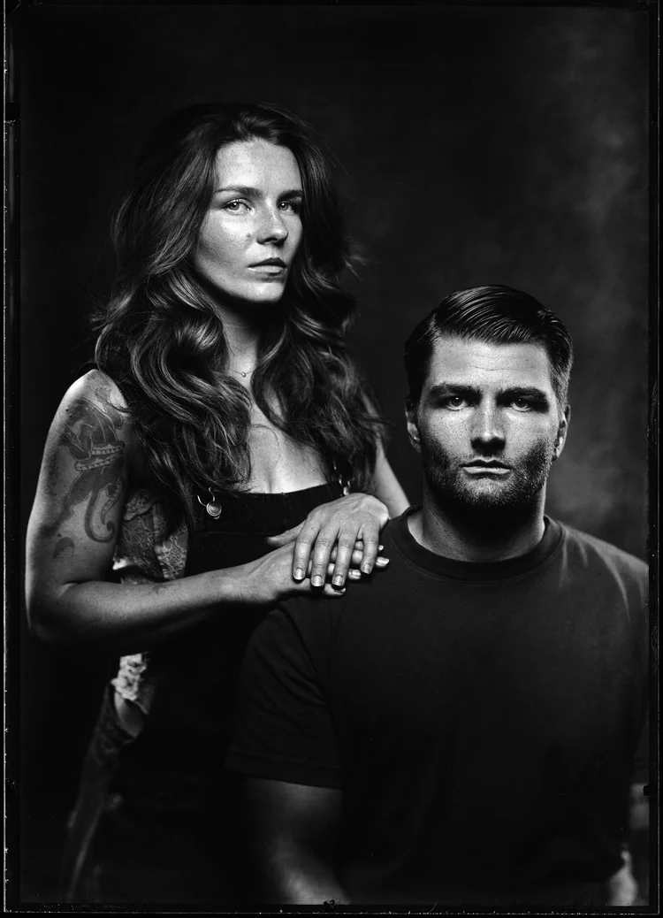 A woman standing behind a seated man, one hand on his shoulder. Her upper arm is tattooed and there are rings on her fingers. Both look straight into the lens. Wet plate collodion tintype.