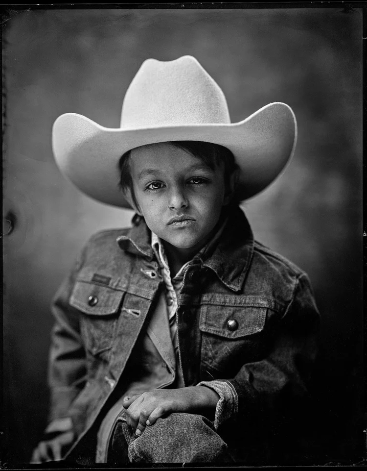 A child under a white cowboy hat several sizes too big, in a denim jacket with their hands folded. The brim takes up most of the frame. Wet plate collodion tintype.