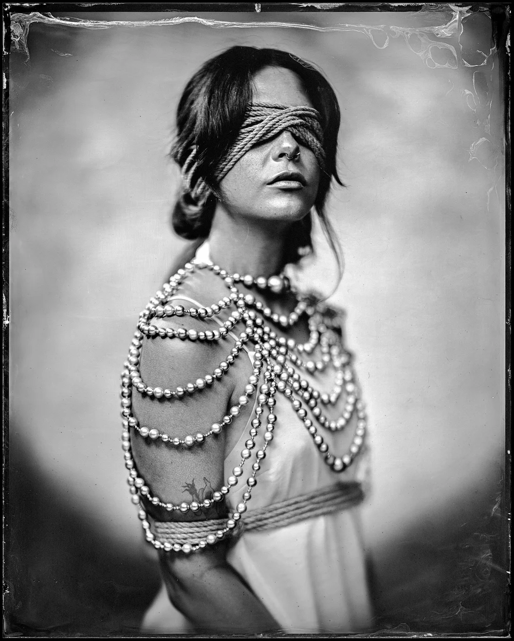 A blindfolded woman in a pale slip, draped in long strands of pearls, with rope tied over her eyes and bound around her arms and waist. Wet plate collodion tintype.