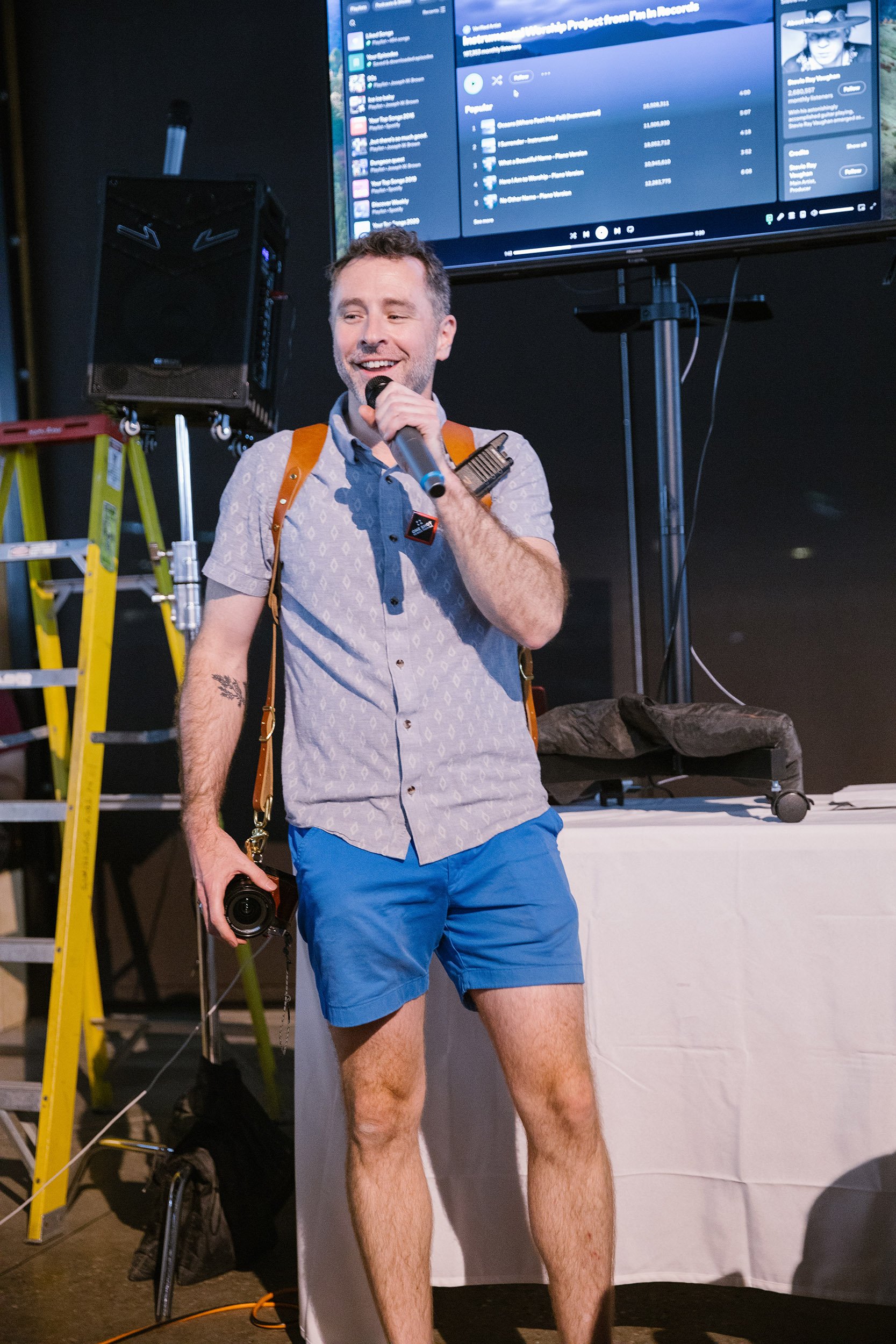 Brett Briggs speaking at an event, a microphone in one hand and a camera in the other