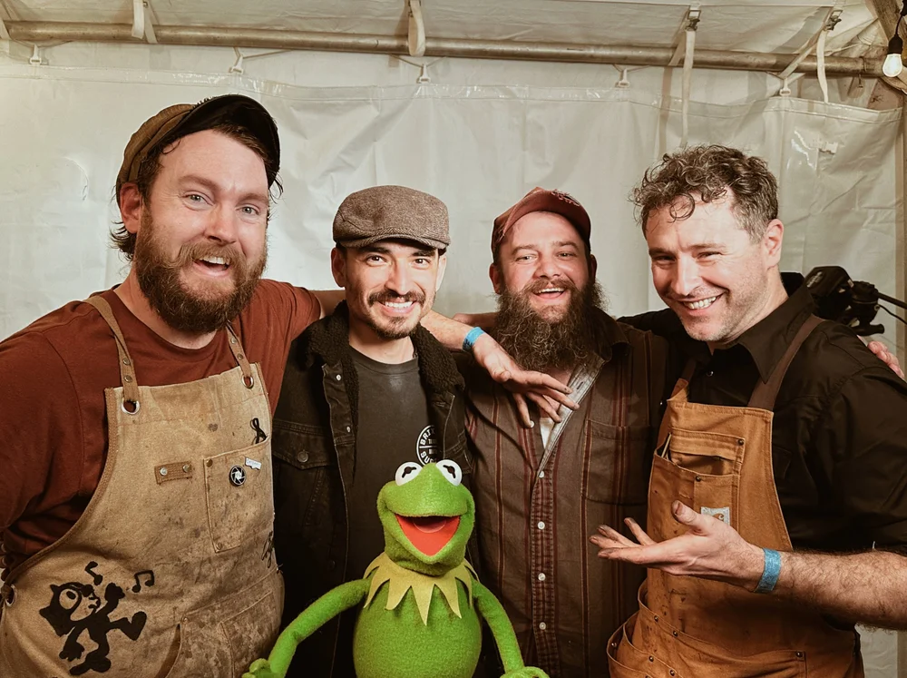 Four men shoulder to shoulder inside a white marquee, grinning at the camera with a Kermit the Frog puppet held up in front of them. Two wear stained canvas work aprons; camera equipment stands at right.