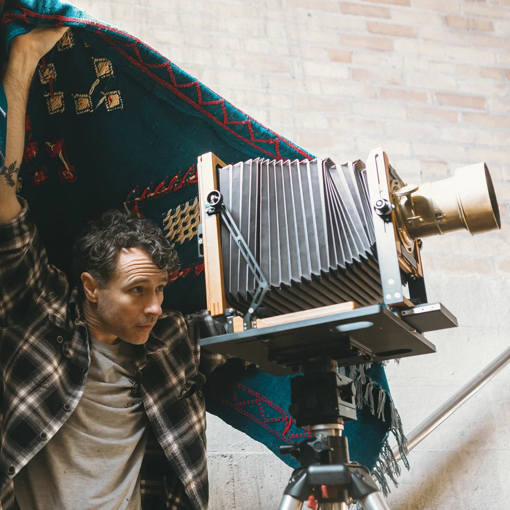 Brett Briggs working under a raised dark cloth beside a large format camera with a brass barrel lens, a patterned dark cloth hung as a backdrop