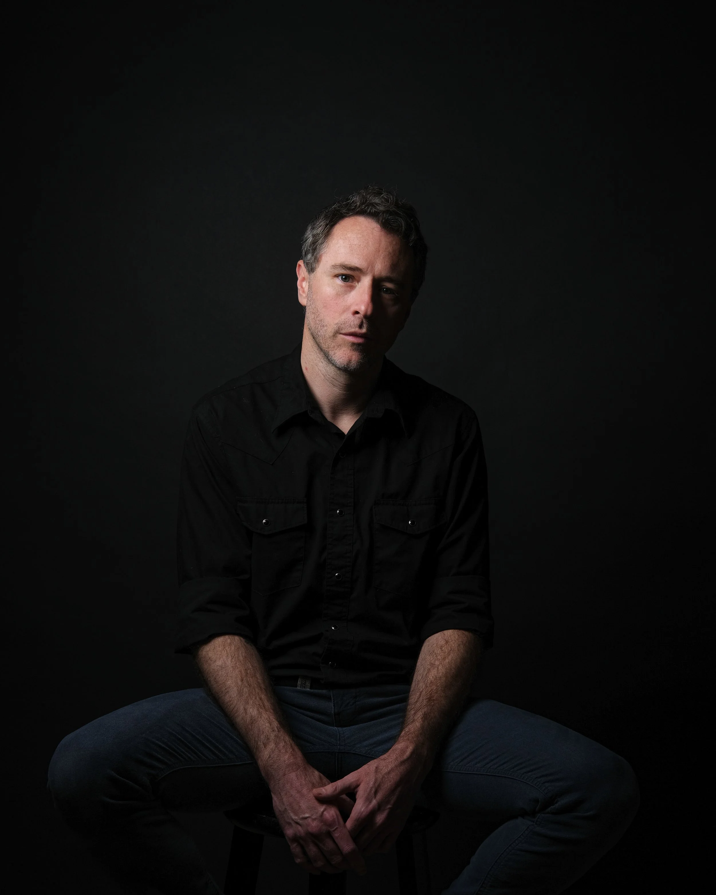 Brett Briggs seated on a stool against a dark background, in a dark button-up shirt and jeans, hands loosely clasped, looking straight into the lens. Photographed by Carlos Guzman.