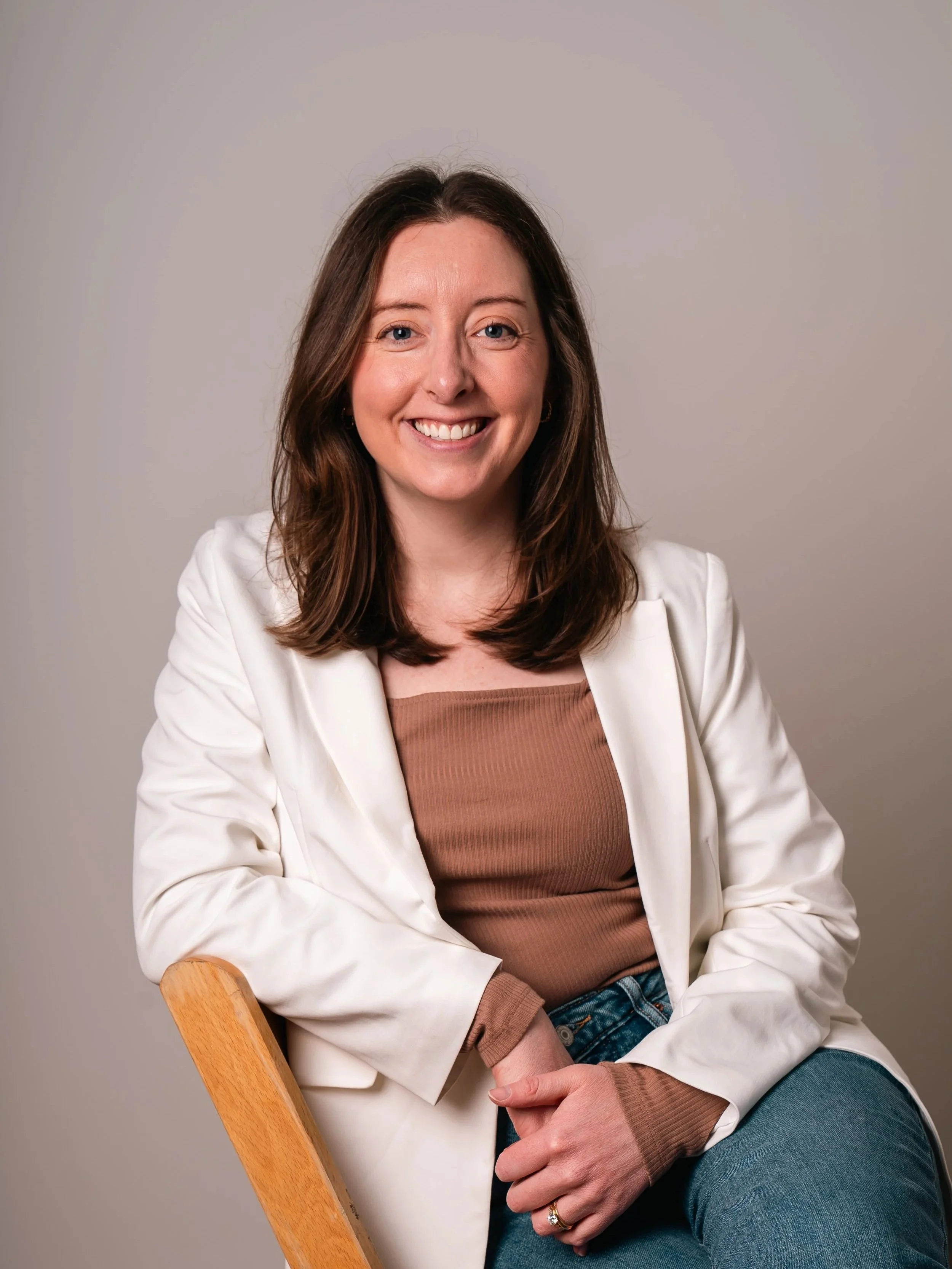 A woman with brown hair, wearing a white blazer, brown top, and jeans, sitting on a wooden chair, smiling at the camera.