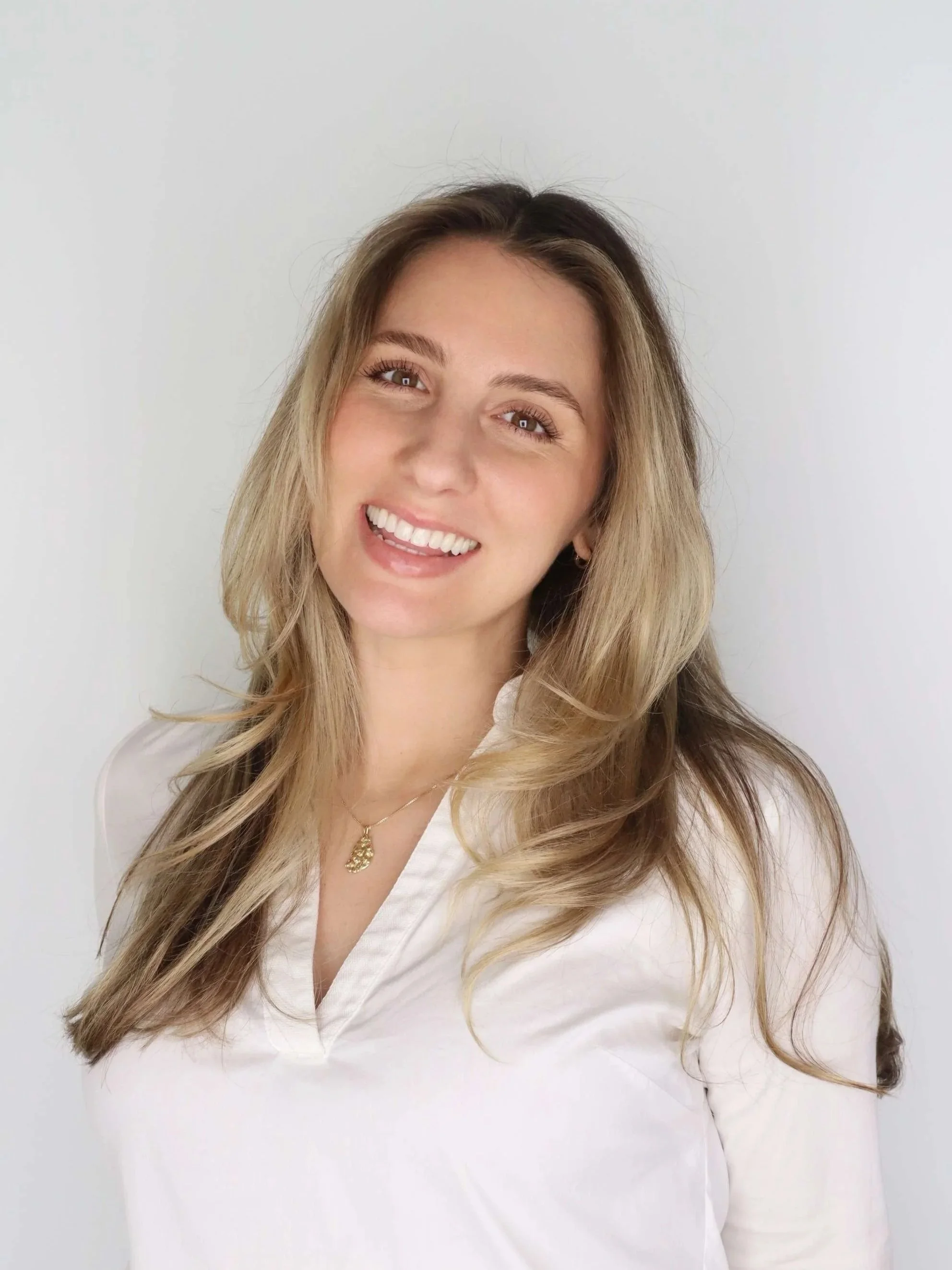 A smiling woman with long, wavy blonde hair wearing a white top and a gold necklace against a plain white background.