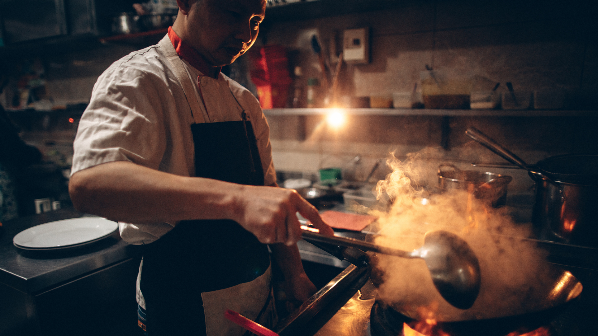 A chef cooking in a professional kitchen with flames rising from a pan, wearing a white chef coat and black apron.