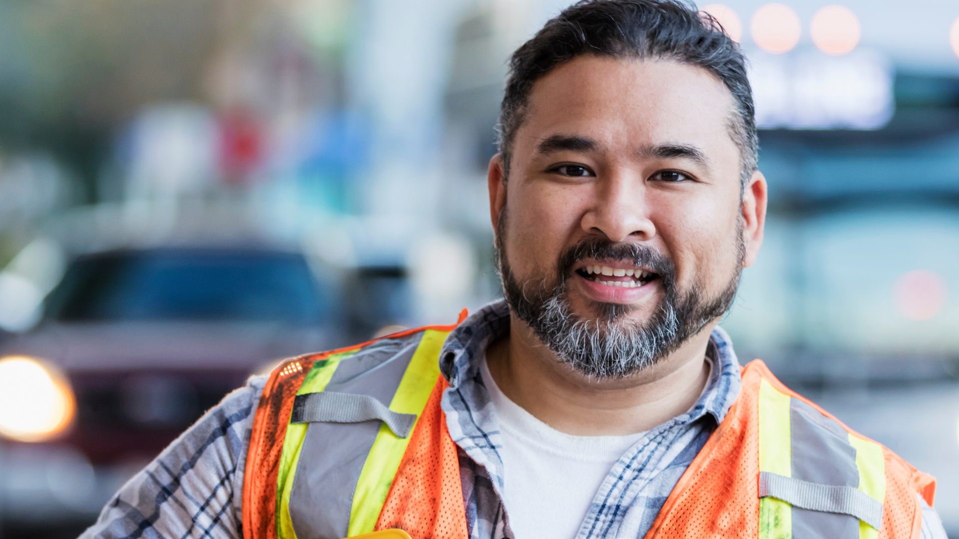 A smiling man wearing a reflective safety vest and a plaid shirt, standing outdoors in an urban area with cars and blurred city lights in the background.