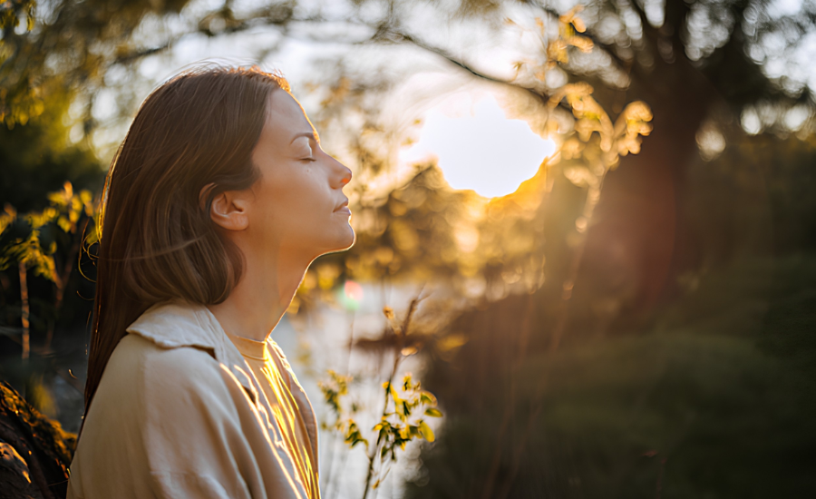 A woman stands outdoors at sunset with her eyes closed, breathing peacefully beside the water as warm golden light filters through the trees.