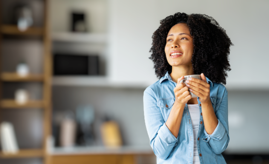A peaceful woman holding a cup of coffee gazes upward with a hopeful expression in a softly lit kitchen.