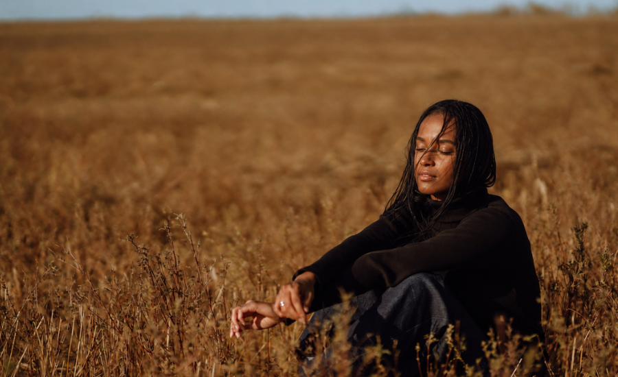 Black woman sitting peacefully in a dry field with her eyes closed, surrounded by golden grass under a clear sky.