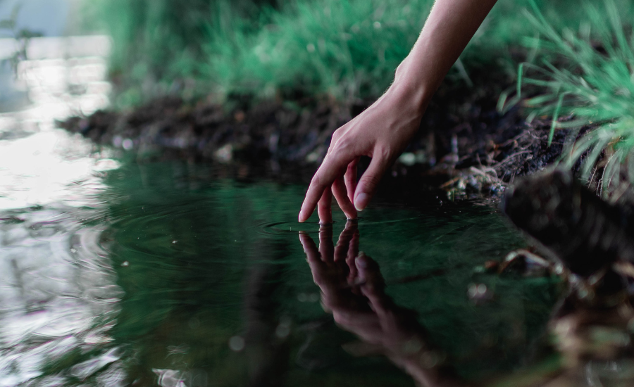 A hand touching still green water at the edge of a bank, ripples spreading outward, symbolizing God's healing touch on bitter waters.