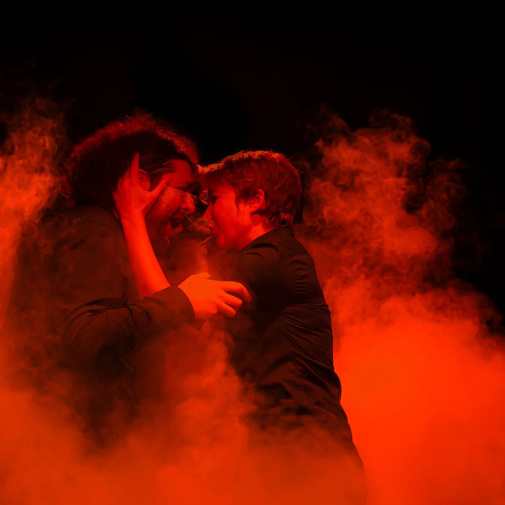 Deux personnes se regardent profondément, entourées de fumée orange et rouge, dans une ambiance sombre et intense.
