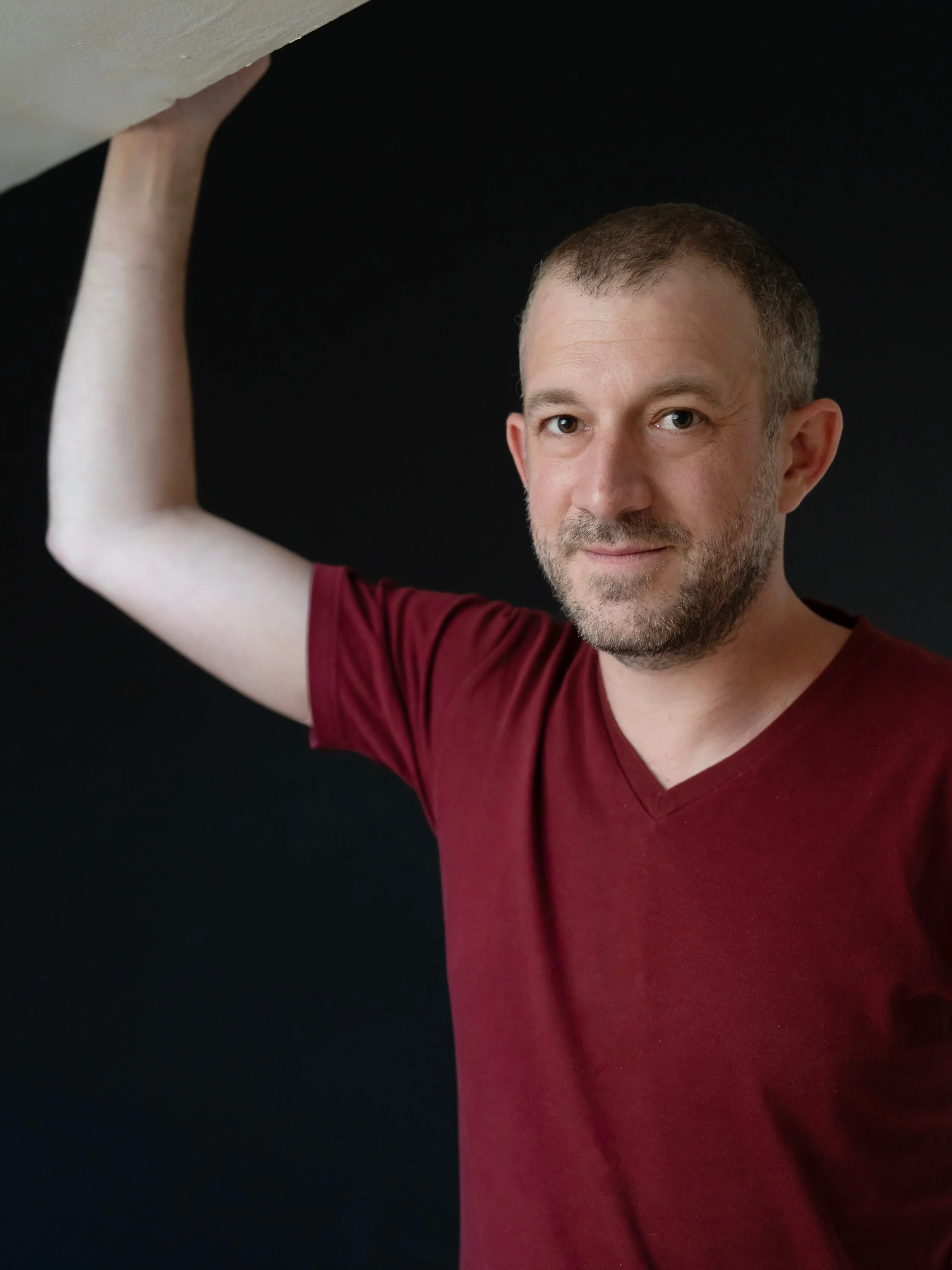 Un homme avec une barbe porte un t-shirt rouge foncé et regarde vers l'appareil photo, tenant un mur ou un objet au-dessus de sa tête contre un fond noir.