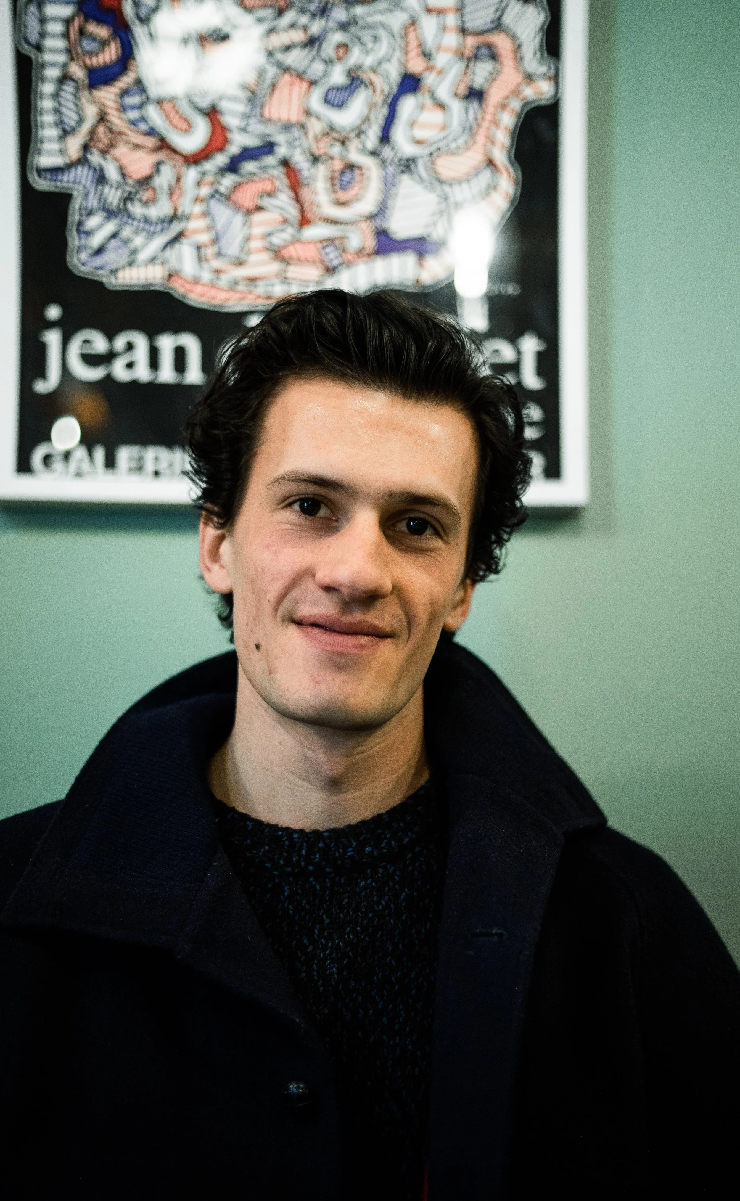 Jeune homme souriant avec des cheveux noirs, portant un manteau noir et un pull gris, debout devant un fond vert avec un tableau contemporain en arrière-plan.