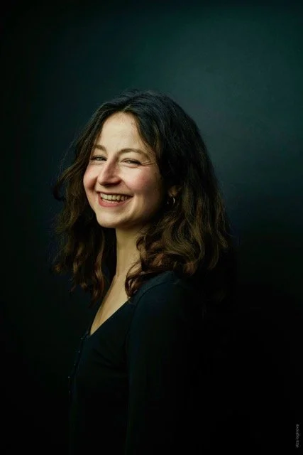 Une femme souriante avec des cheveux bruns et bouclés, portant un haut noir, contre un fond noir.
