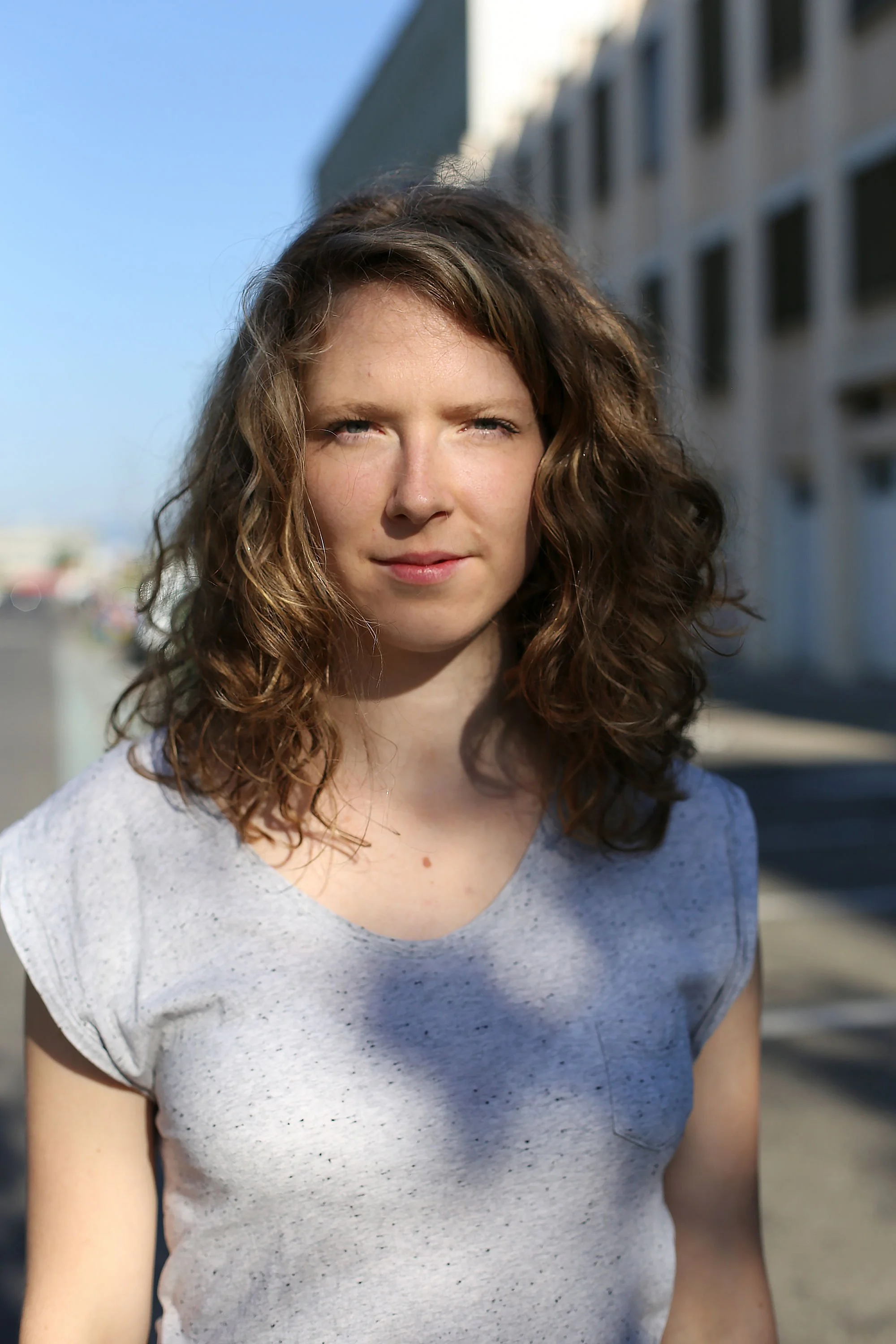 Jeune femme aux cheveux bouclés bruns portant un t-shirt gris clair, t-shirt à manches courtes, dans un décor urbain avec bâtiment en arrière-plan