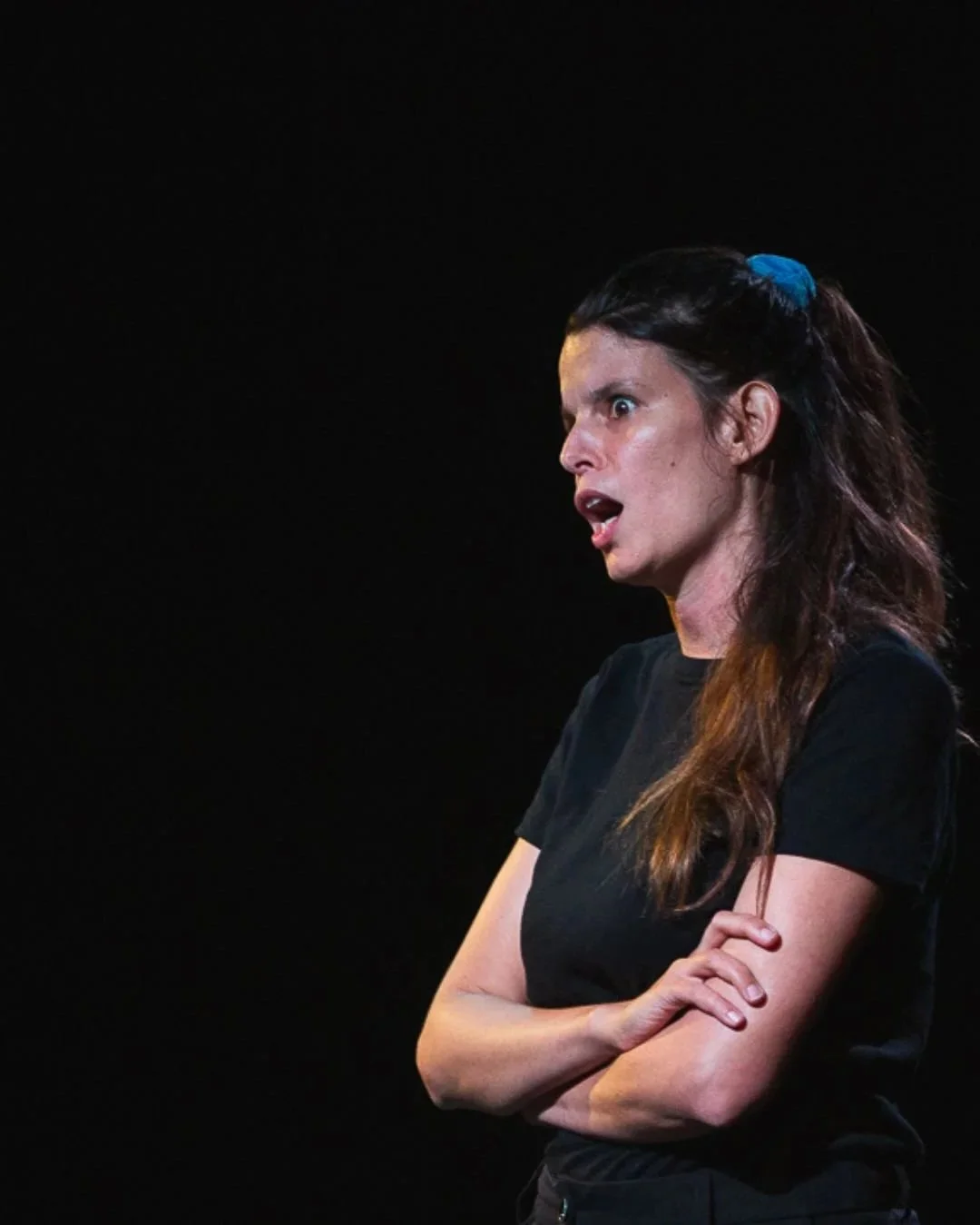 Une femme avec des cheveux bruns et un ruban bleu dans les cheveux, portant un t-shirt noir, semble exprimer la surprise ou la colère, avec ses bras croisés et un fond noir.
