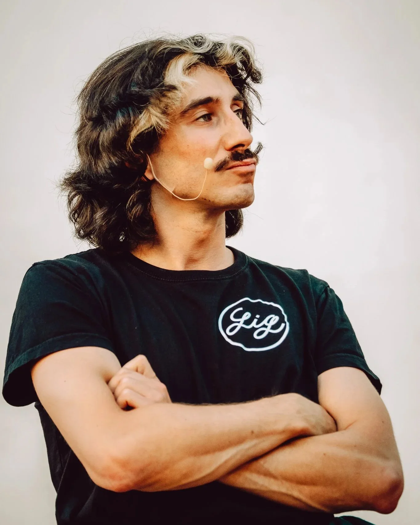 Un homme avec des cheveux longs et bouclés, portant un t-shirt noir avec un logo blanc, croise les bras et regarde vers la droite.