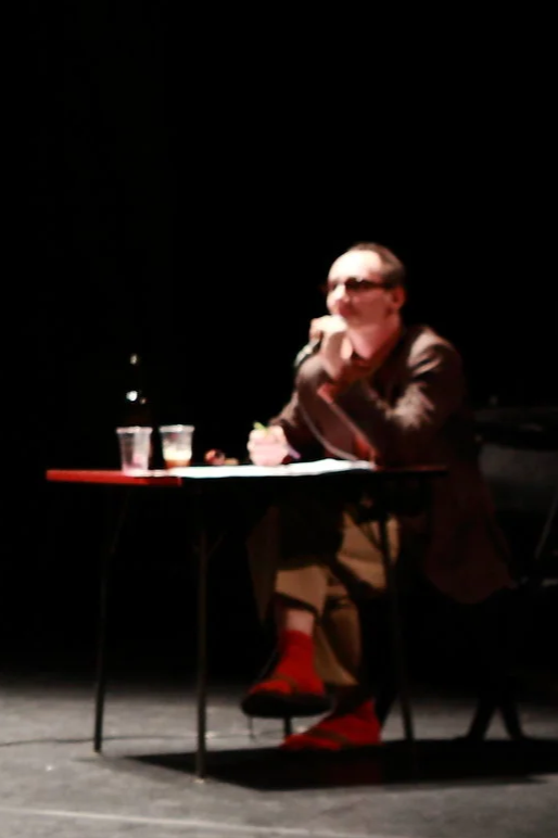 Personne assise à une table sur scène, avec deux verres d'eau, portant un costume avec des chaussettes rouges, sous un éclairage tamisé.