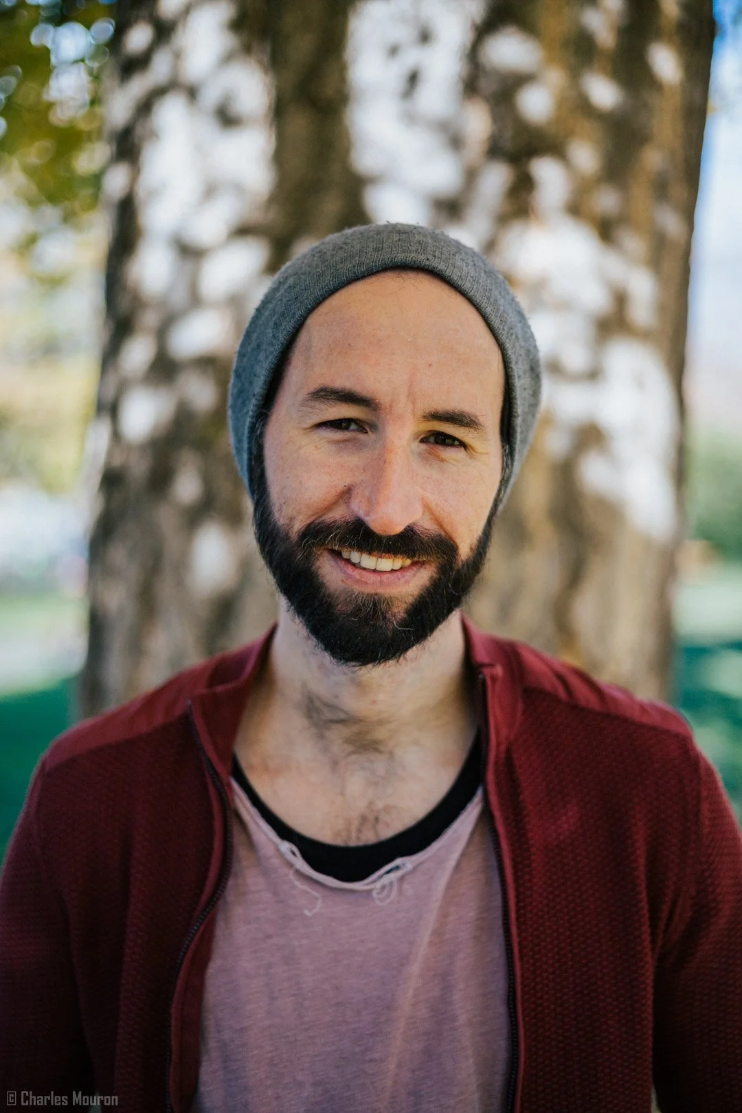 Un homme souriant avec une barbe portant un bonnet gris, une veste rouge et un t-shirt rose, devant un arbre. Environnement extérieur, journée ensoleillée.